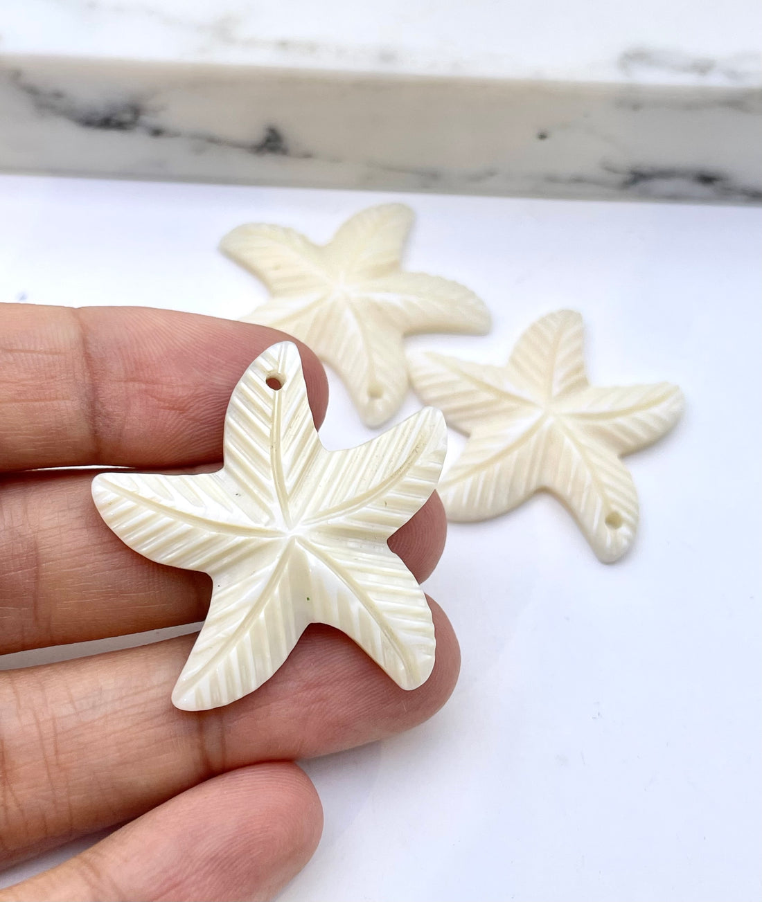 A hand holds a carved starfish-shaped pendant in the foreground, with two similar starfish pendants blurred in the background on a white surface.