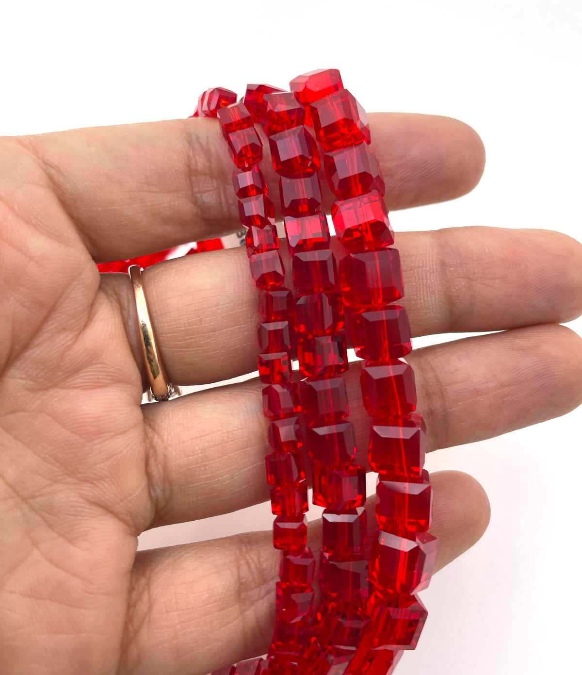 A hand holding three strands of faceted red glass beads, with a gold ring visible on one finger, against a plain white background.
