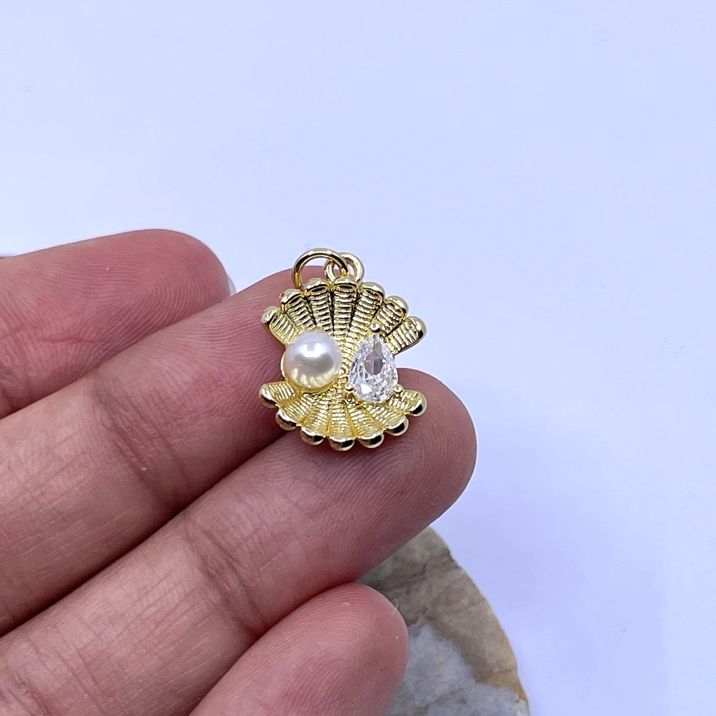 A small gold shell-shaped charm with a pearl and clear teardrop gemstone, held between fingers against a pale blue background.