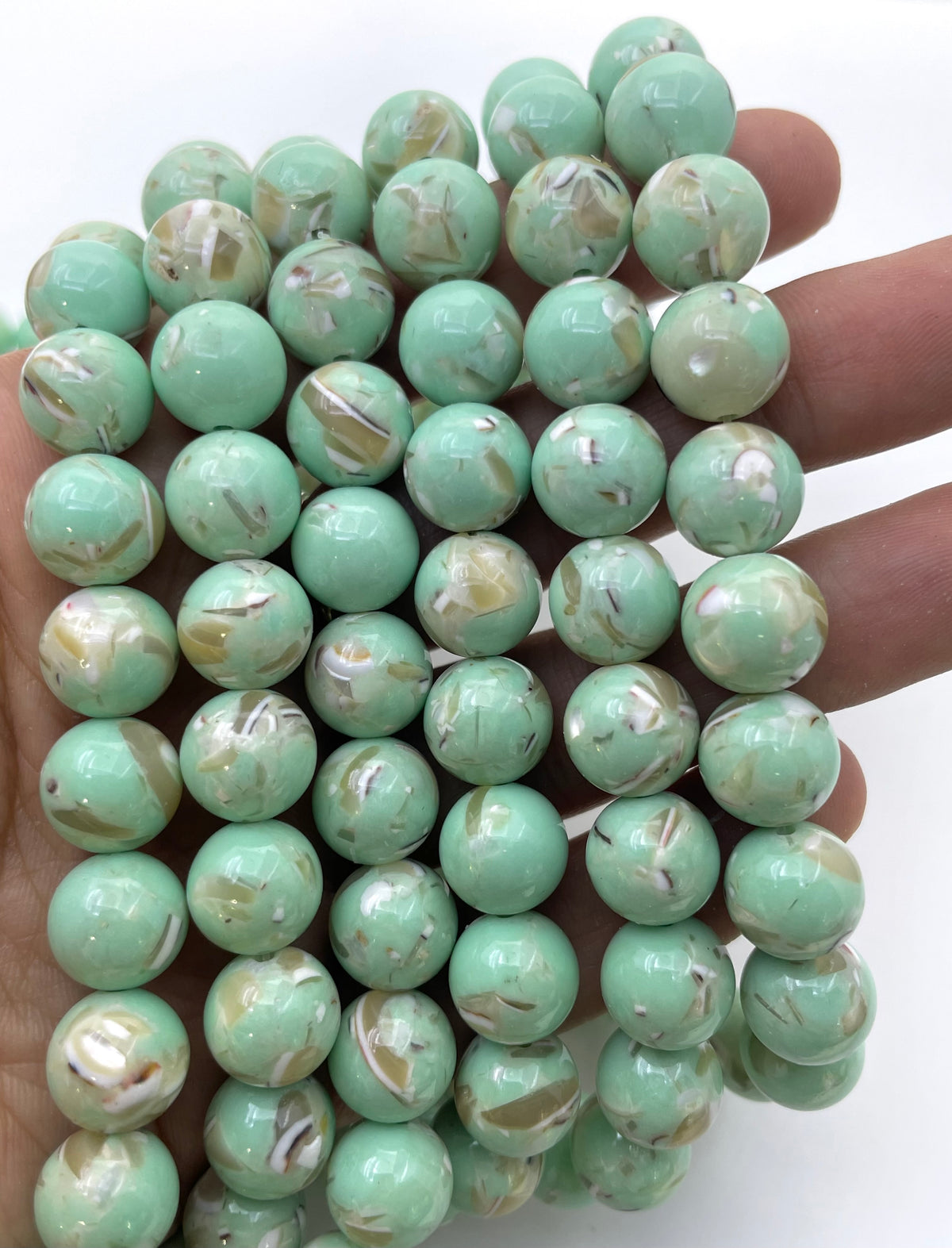 A hand holding a bundle of round, glossy mop beads in a light mint green color with white and beige marbled swirls, set against a plain white background.
