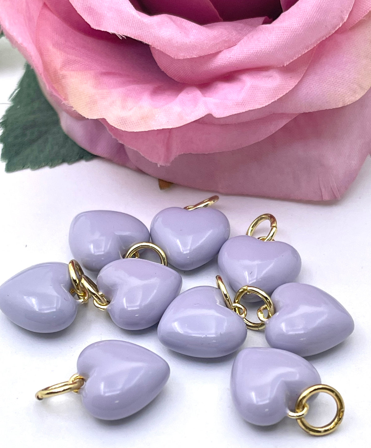 Ten lavender heart-shaped charms with gold-colored metal loops, arranged on a white surface with a pink rose in the background.
