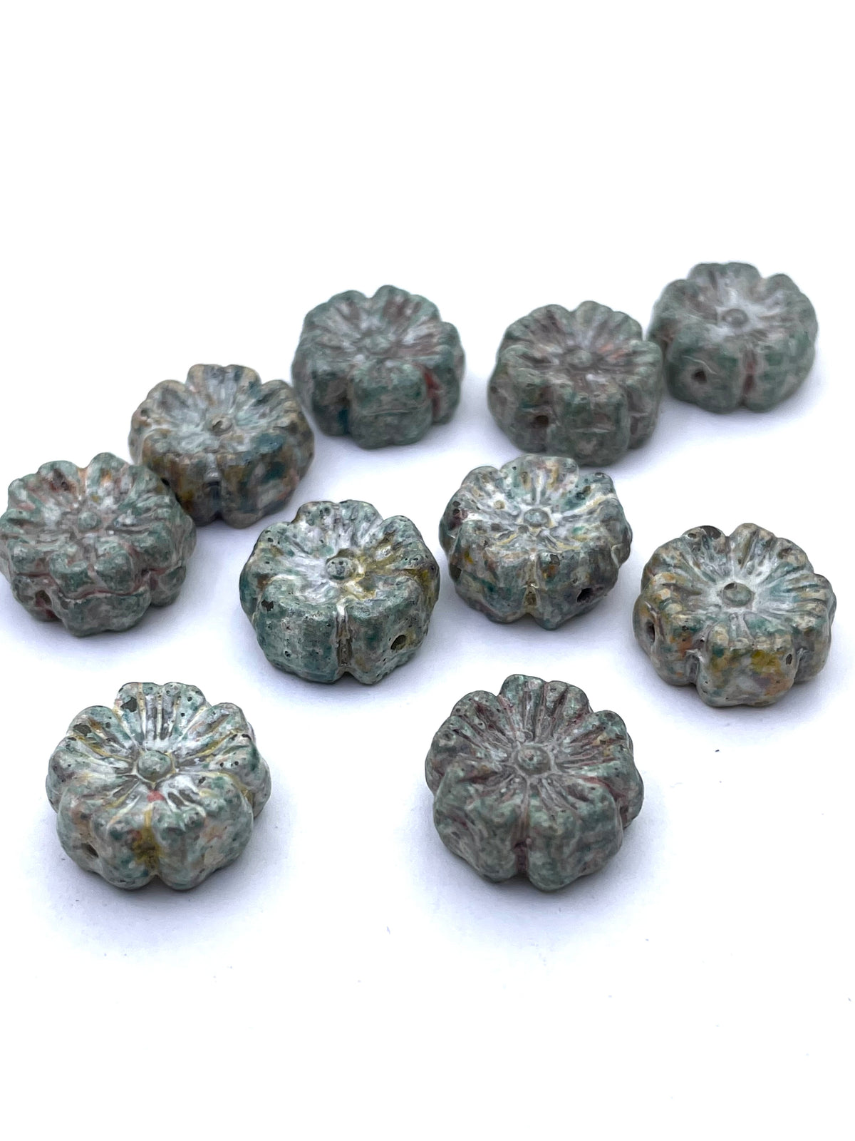 Nine small flower-shaped beads with a distressed, textured finish in muted green, gray, and brown tones, arranged on a plain white background.