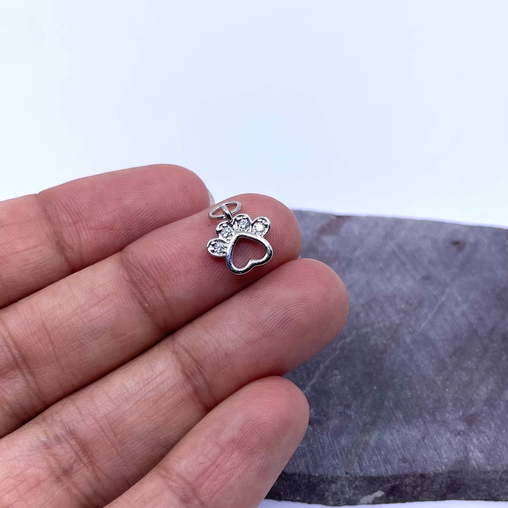 A silver paw print charm held on a person's fingers to show its small scale, with a gray stone background.