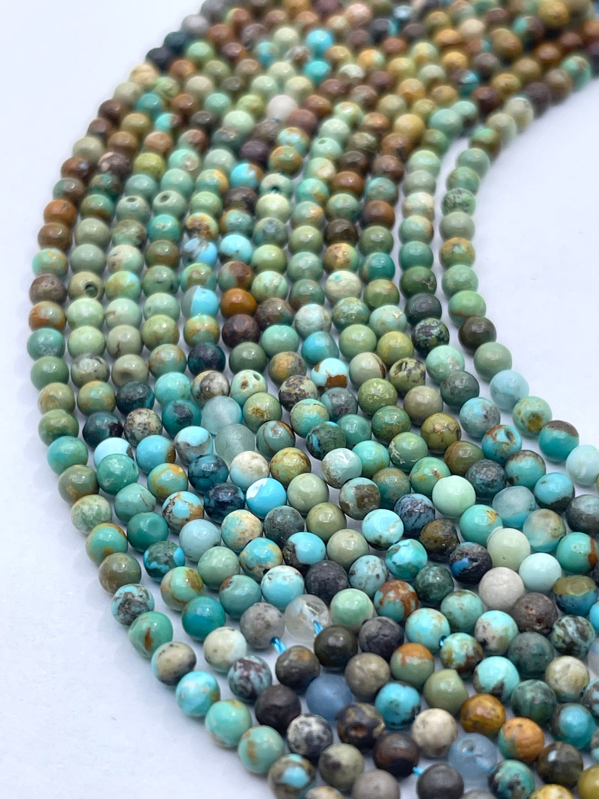 Multiple strands of round turquoise beads with natural marbled patterns in shades of blue, green, brown, and white, arranged on a plain white background.