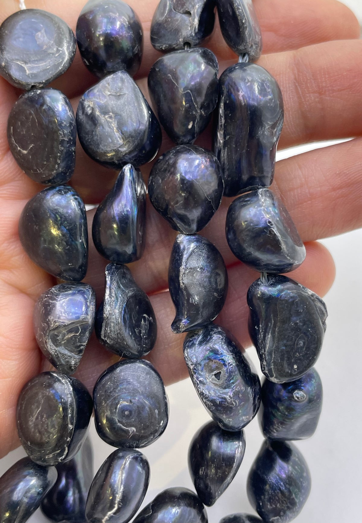 A close-up of a hand holding strands of polished, irregular dark gray to black baroque pearls or stone beads with a subtle iridescent sheen.