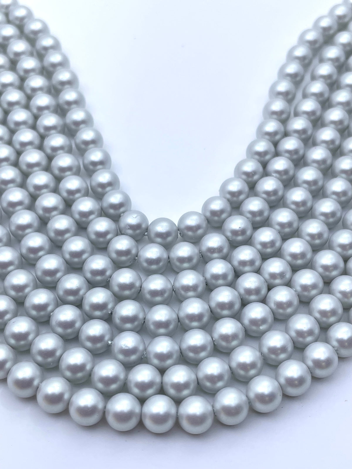 Multiple strands of round, iridescent shell pearl beads in a light silver-gray color, arranged in a V-shape on a plain white background.