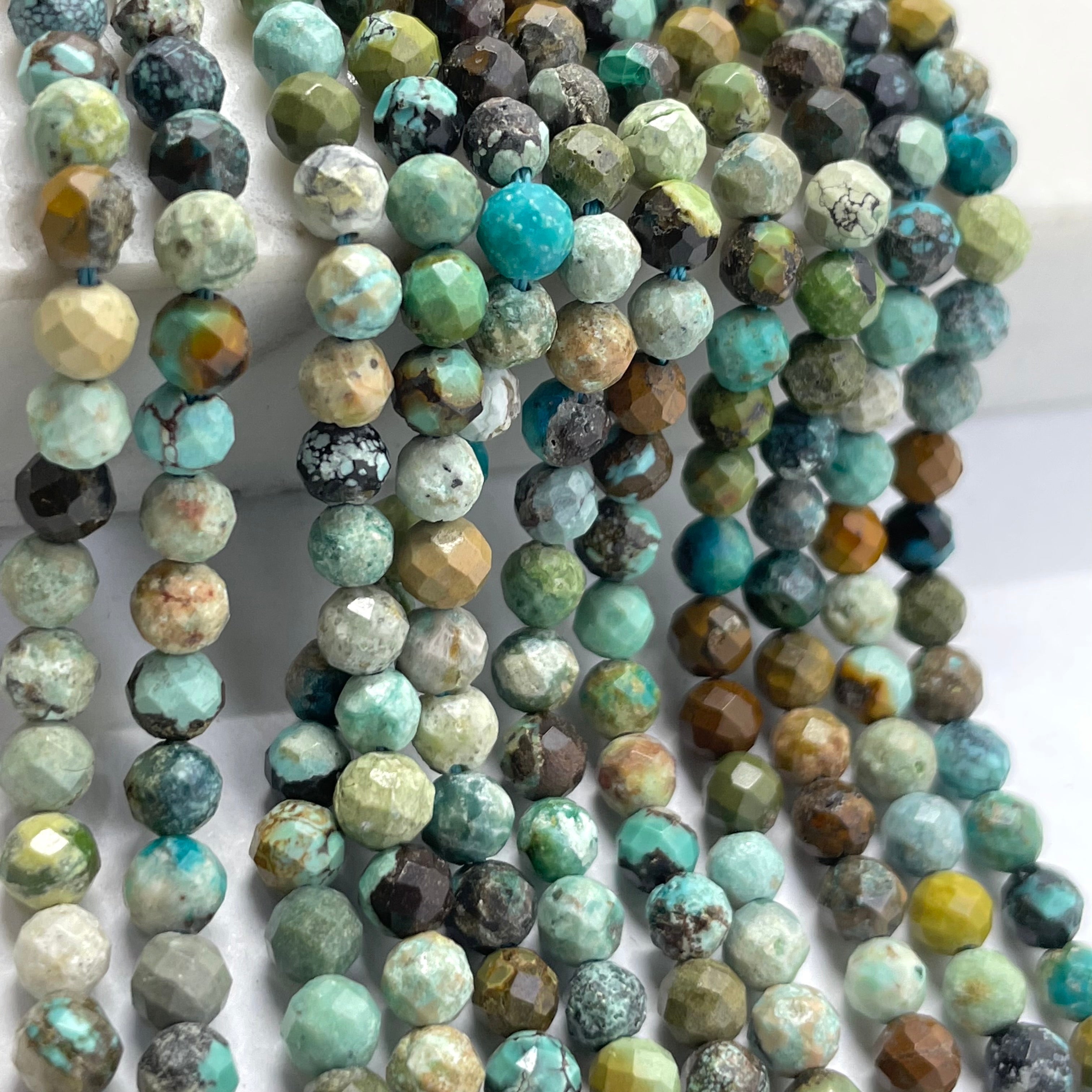 Multiple strands of faceted round turquoise beads in a marbled mix of turquoise, green, brown, gray, and cream colors, draped over a white surface.