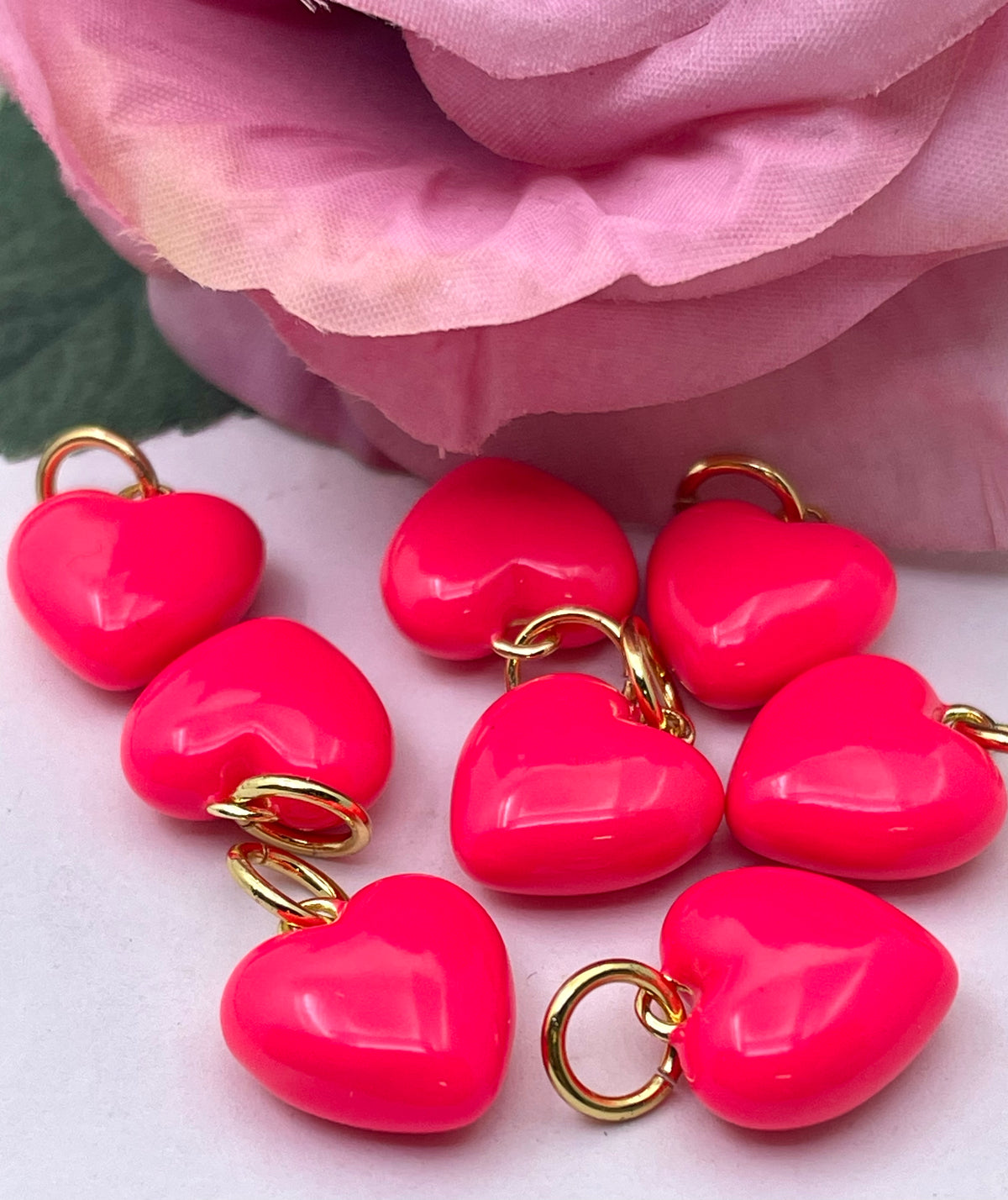 Several glossy hot pink heart-shaped charms with gold-tone split rings, arranged on a white surface with a soft pink fabric rose in the background.