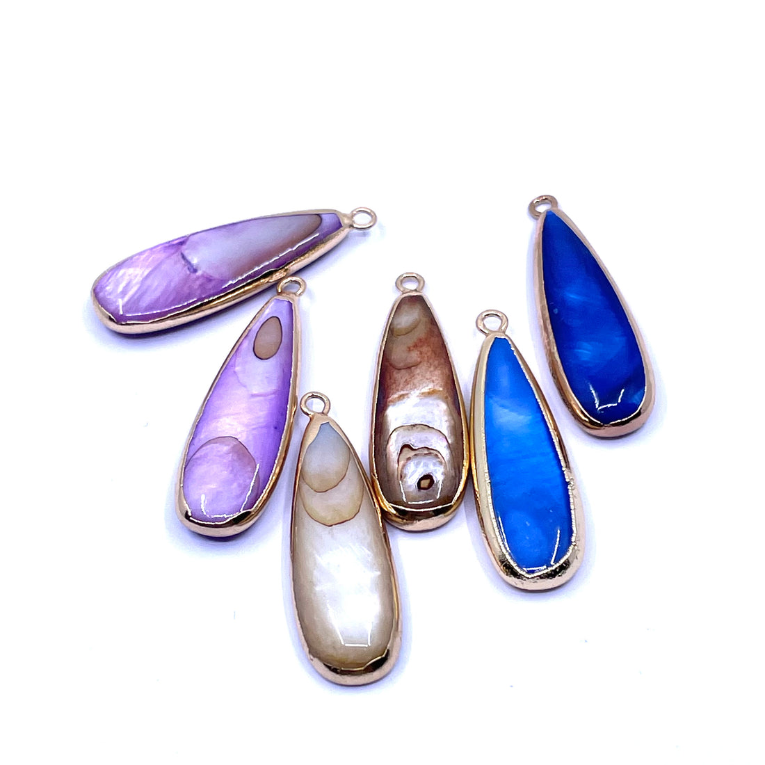Shell pendants in gold-tone bezel settings, displayed on a clean white background. The stones feature various colors and patterns, including shades of purple, blue and brown.