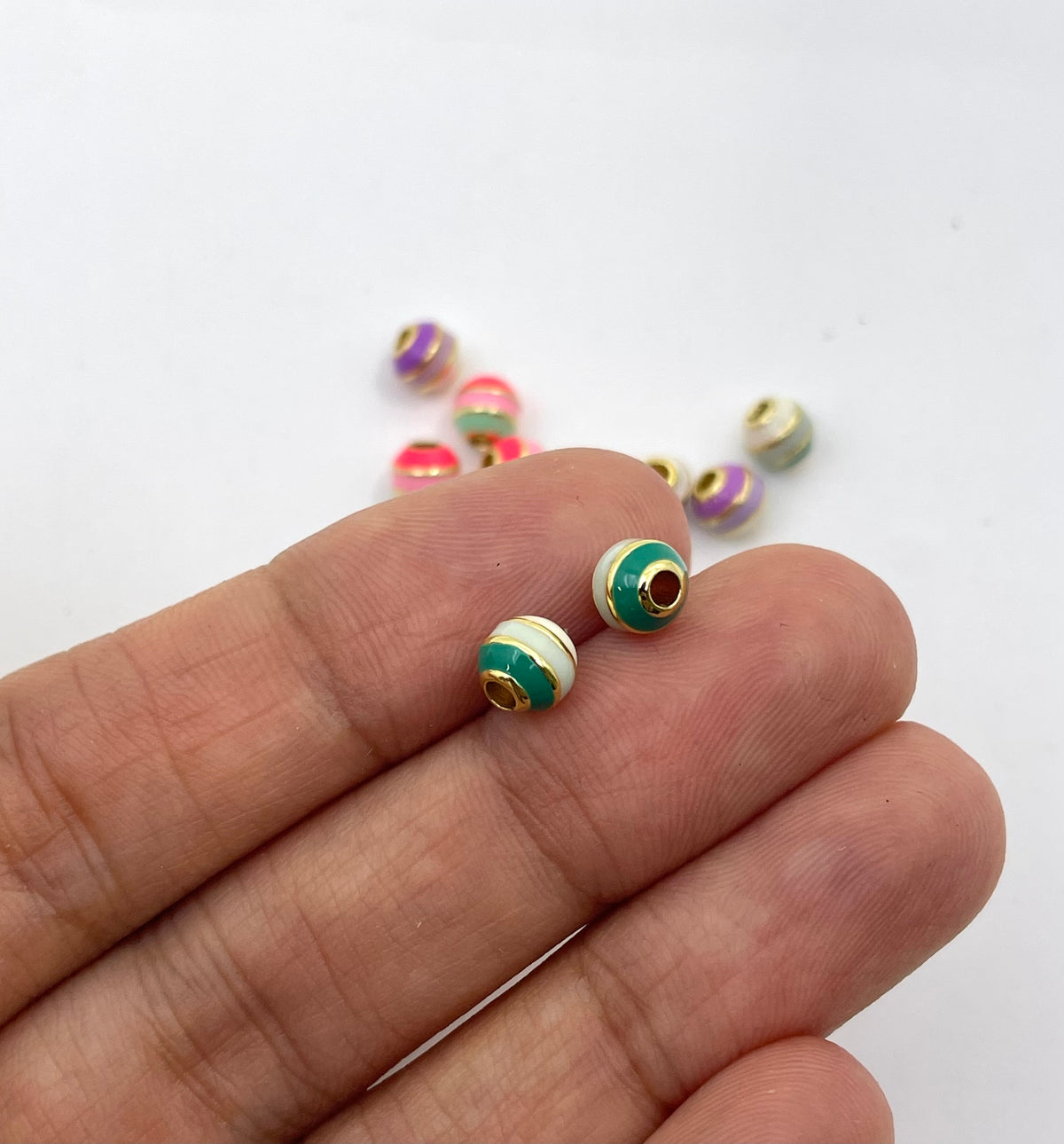 A close-up of a person's hand holding two small striped beads with green, white, and gold accents, with more similar beads scattered in the background on a white surface.