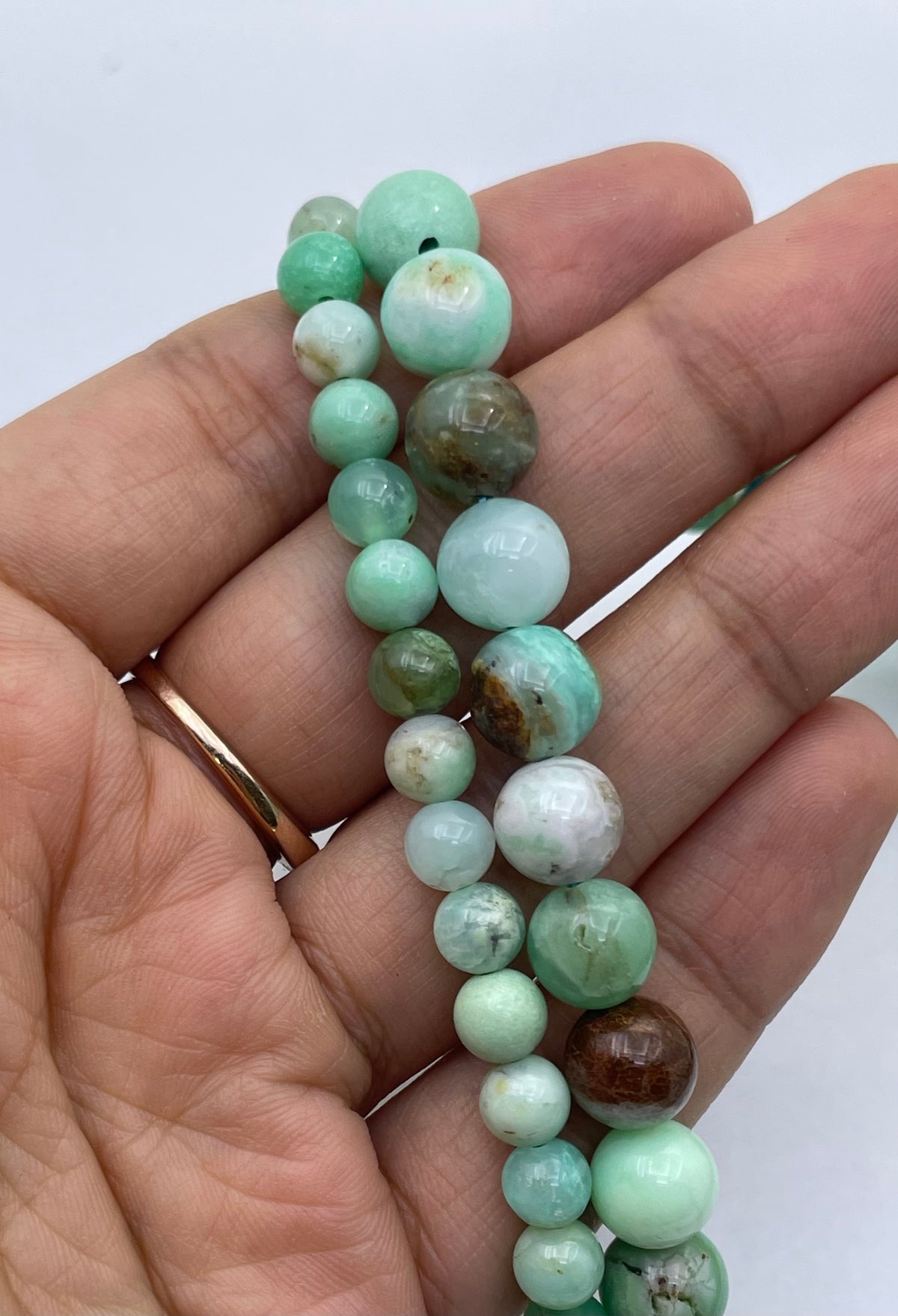 A close-up of a hand holding a strand of polished round beads in mint green, white, and brown marbled stone, with a gold ring on the finger.