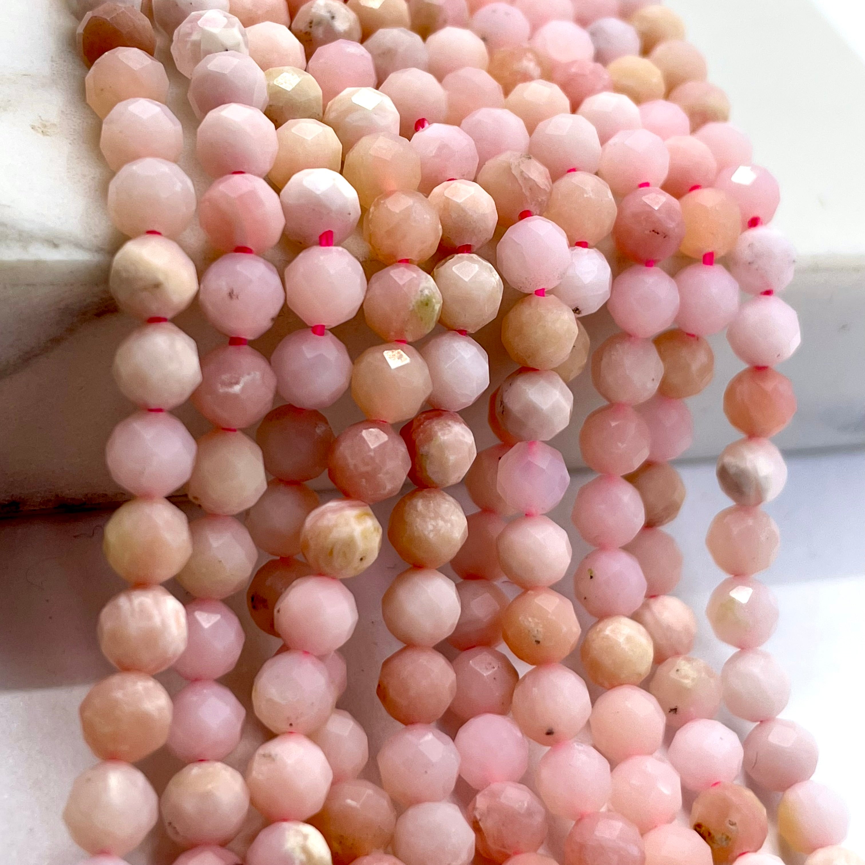 Multiple strands of faceted round pink opal beads in soft pink, peach, and cream tones draped over a white marble display block.