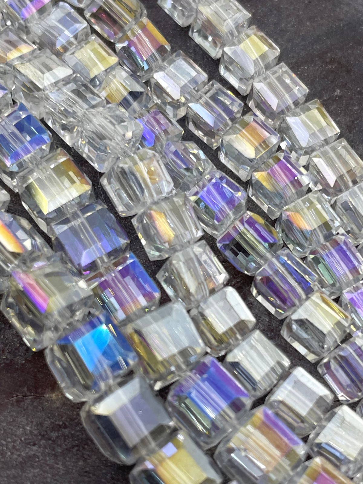 Clear glass AB beads with an iridescent rainbow finish, arranged in diagonal rows on a dark textured background.