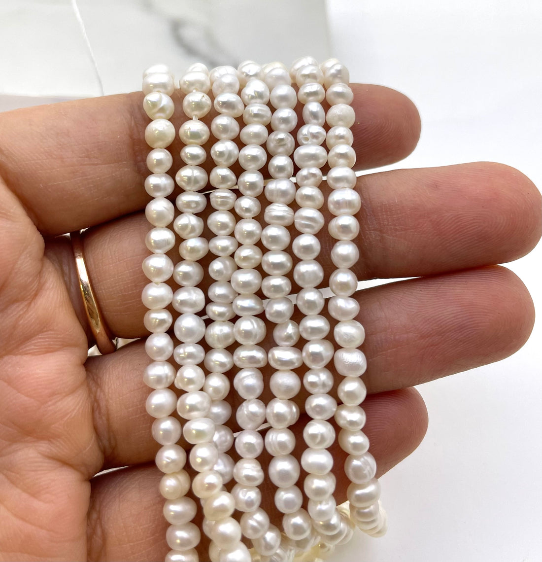 A hand holding several strands of white freshwater pearls, with a gold ring visible on the ring finger, against a light background.