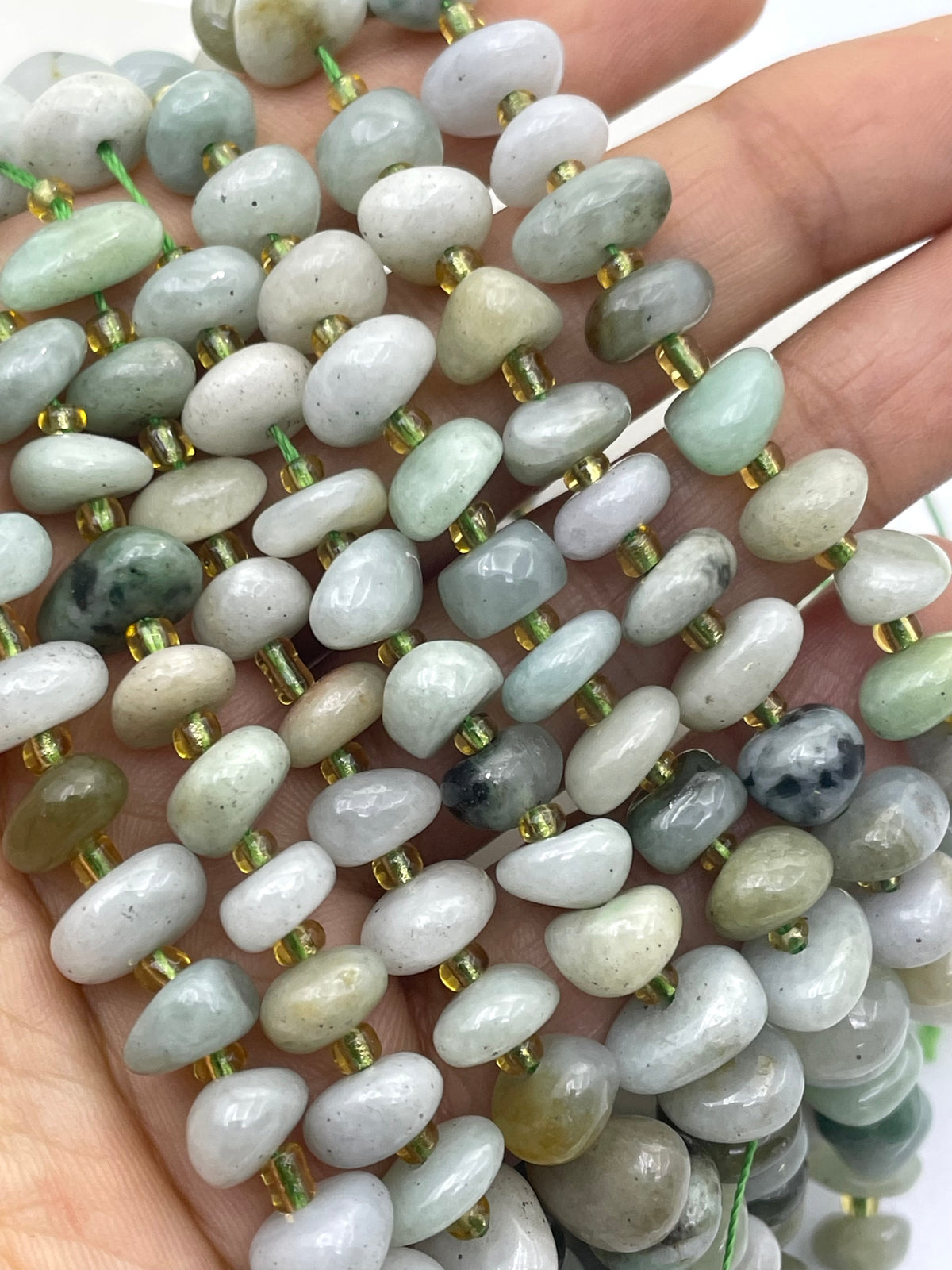 A close-up of a hand holding multiple strands of polished burma jade stone beads threaded on gold-tone cords. The beads are smooth, rounded, and come in soft pastel shades of pale green, gray, white, and some darker mottled areas.