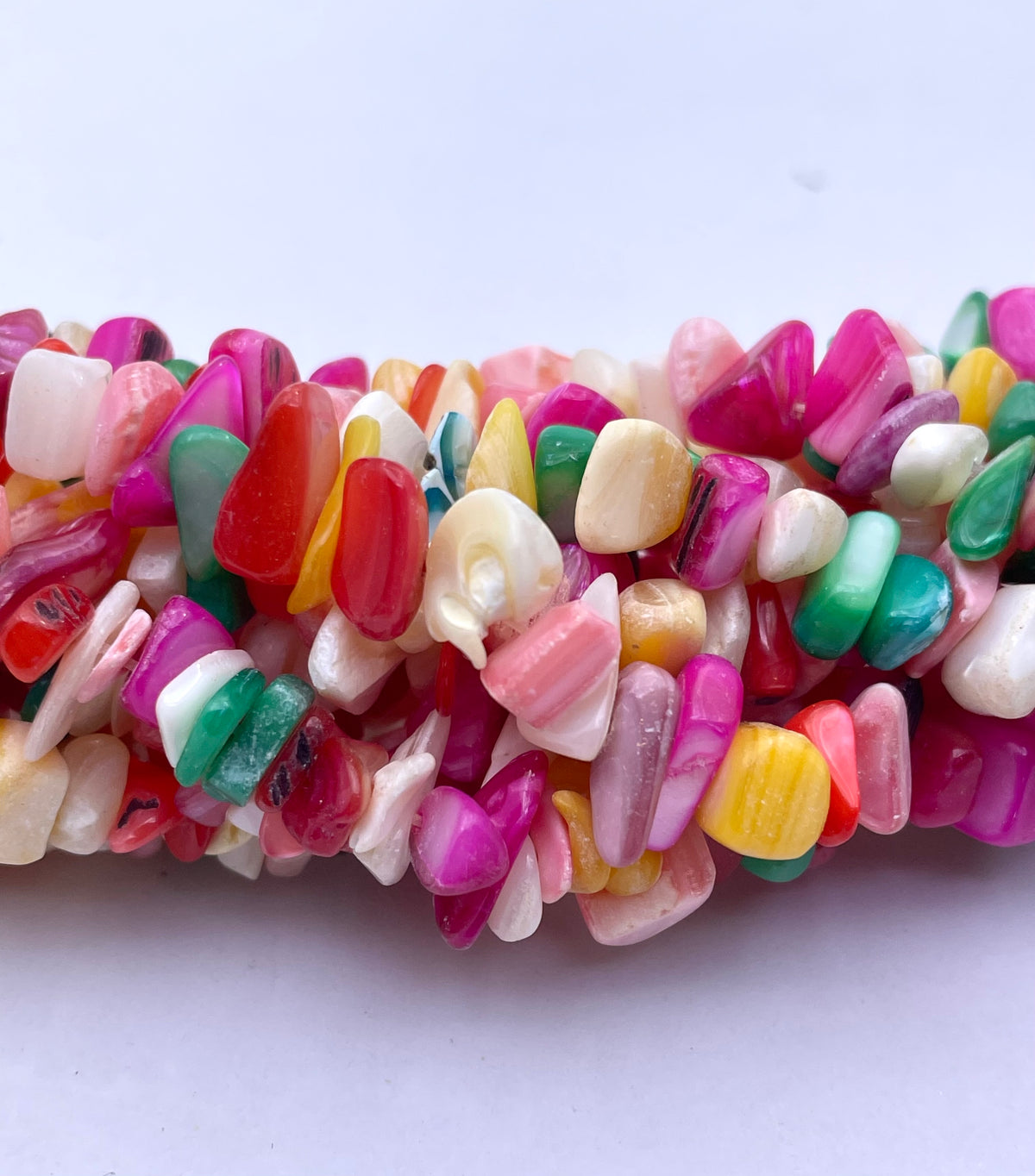 A close-up of a strand of polished tumbled mop chips in a variety of bright colors including pink, red, green, yellow, white, and orange, arranged horizontally on a plain light background.