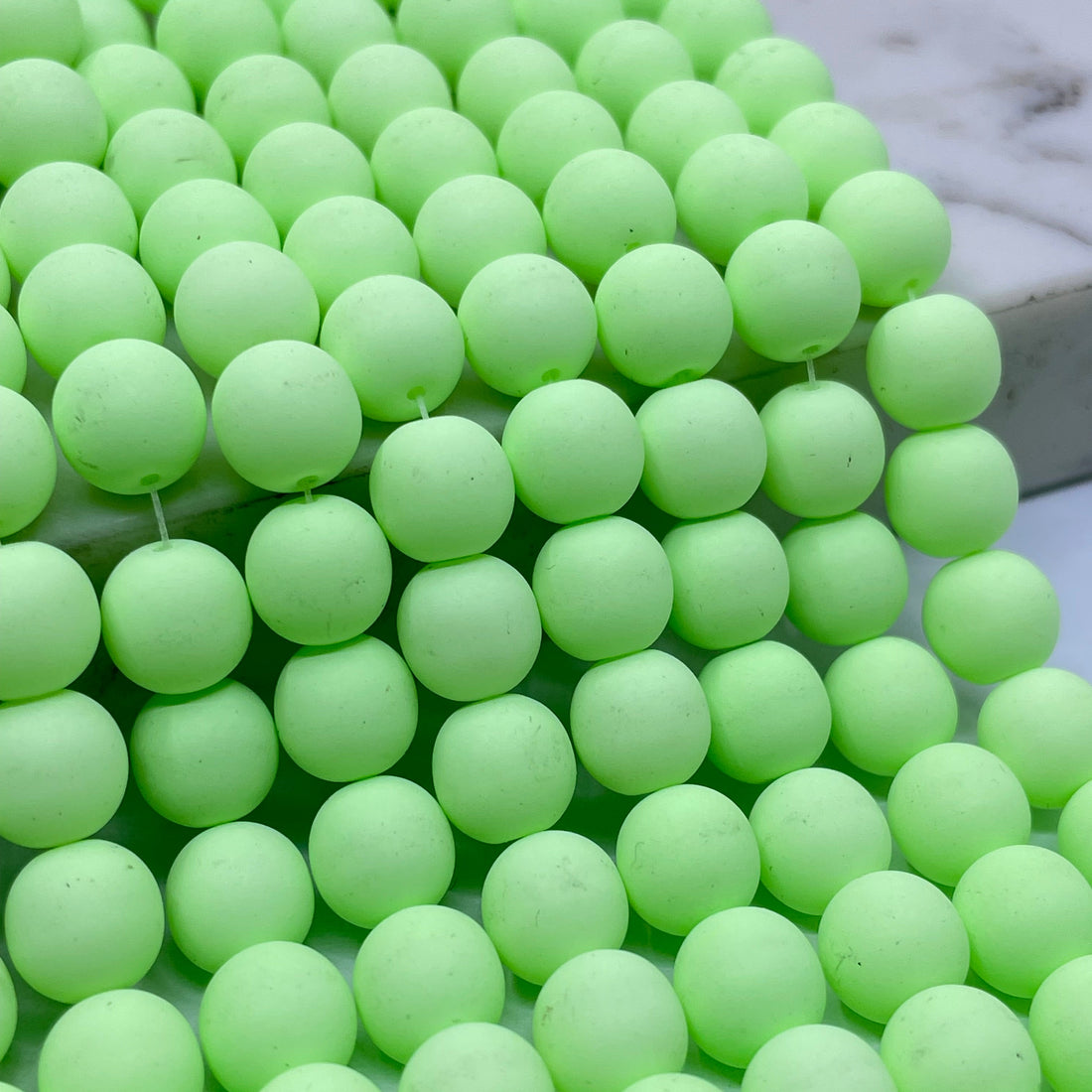 A close-up of many pale green grape-like beads called velvet beads or pom-pom balls arranged in rows, with a small white marble-like surface visible in the upper left corner.