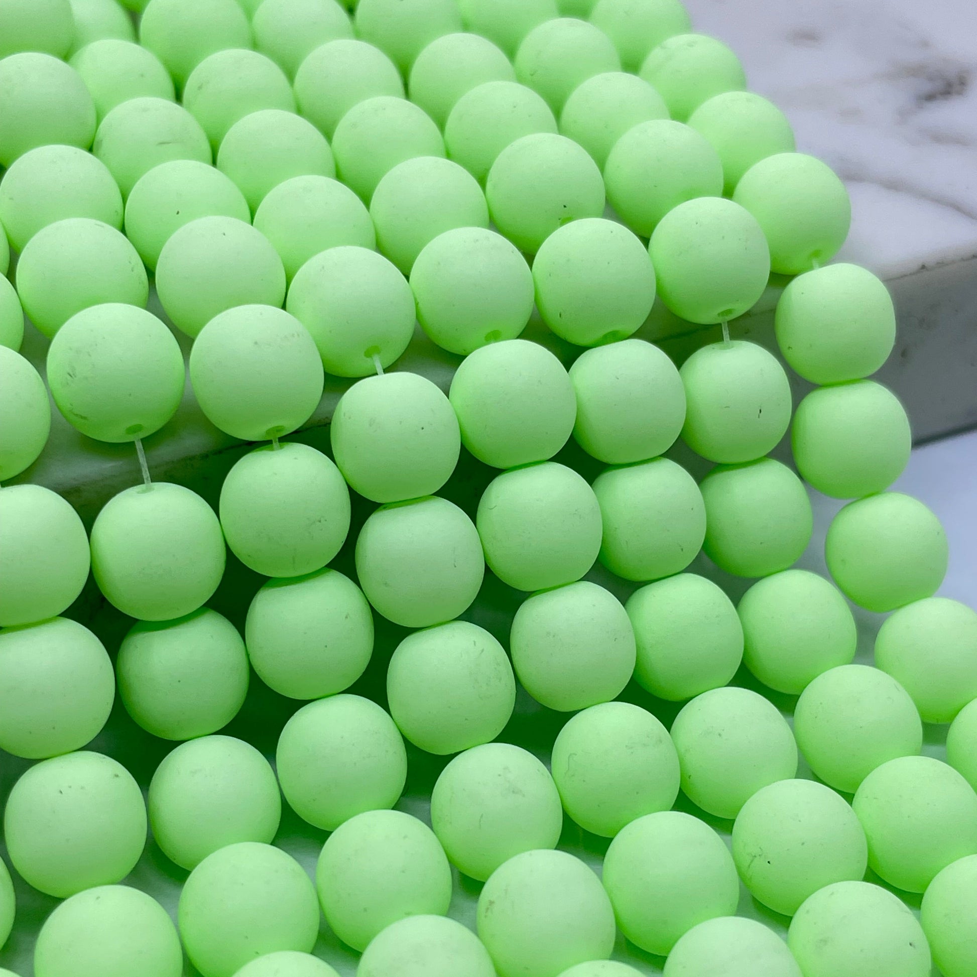 A close-up of many pale green grape-like beads called velvet beads or pom-pom balls arranged in rows, with a small white marble-like surface visible in the upper left corner.