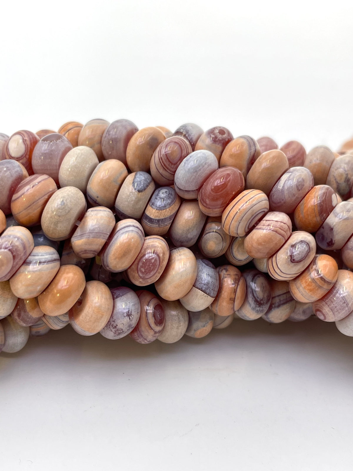 A close-up of multiple strands of polished gobi agate stone beads in earthy neutral tones, including beige, brown, gray, and muted purple, arranged horizontally against a plain white background.