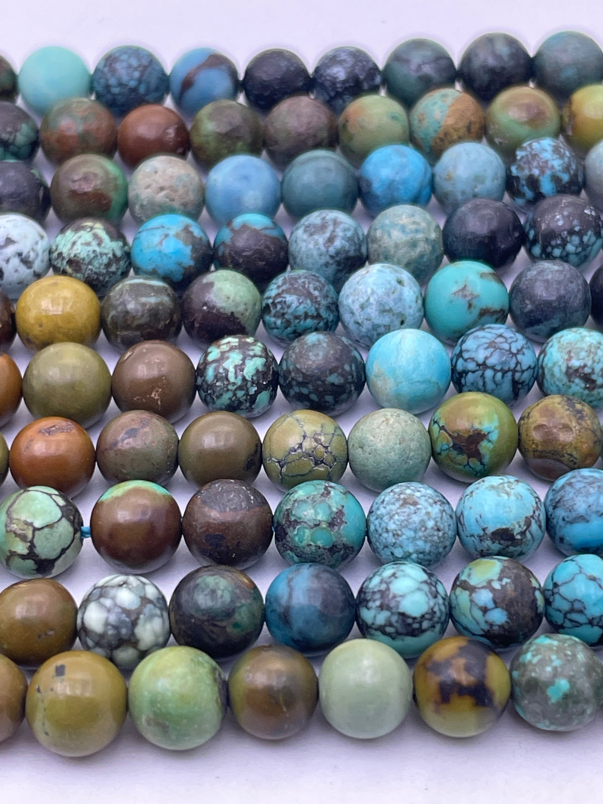 Multiple strands of round turquoise stone beads arranged in rows on a plain white background. The beads display natural marbled patterns in shades of blue, green, brown, and gray.