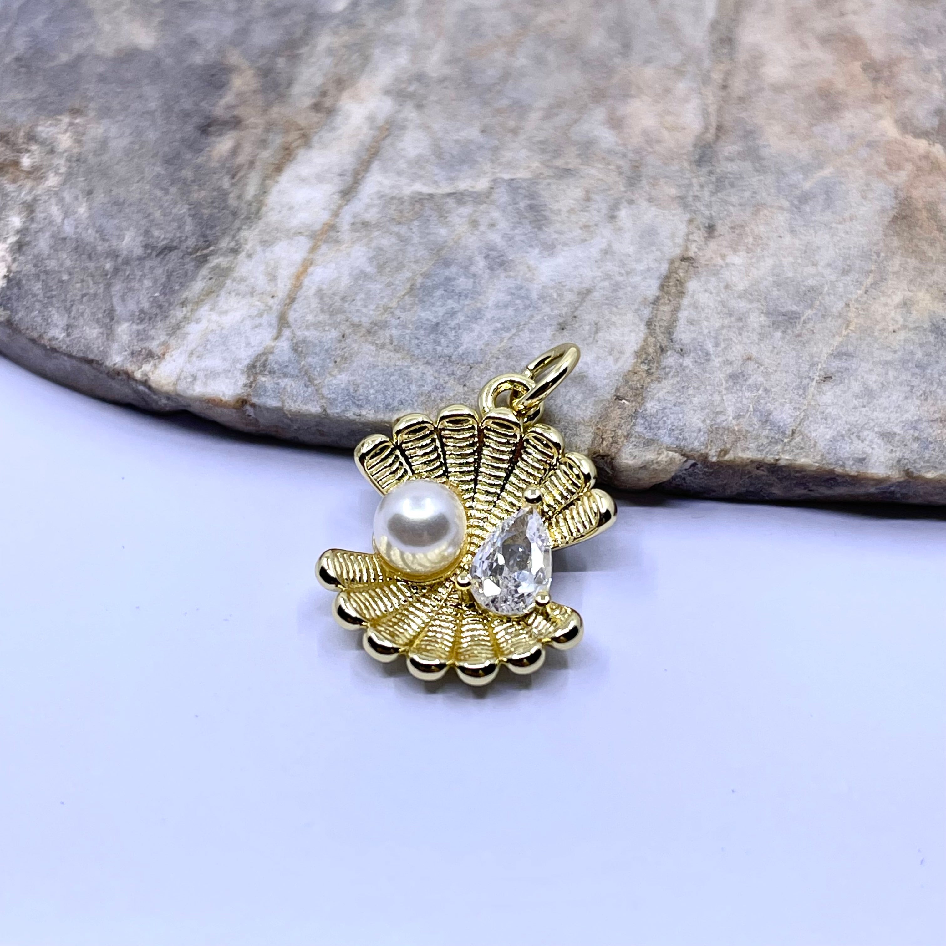 A gold-toned shell-shaped pendant charm featuring a white pearl and a clear teardrop crystal, displayed on a light surface with a gray stone in the background.