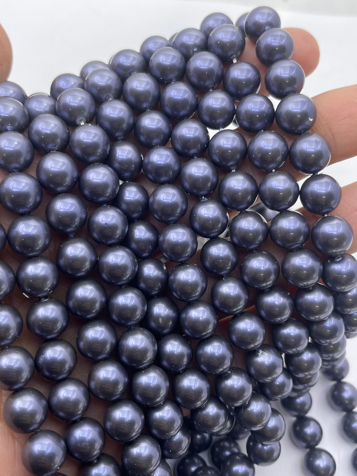 A close-up of a hand holding a strand of dark blue-gray, shell pearl beads with a subtle iridescent sheen.