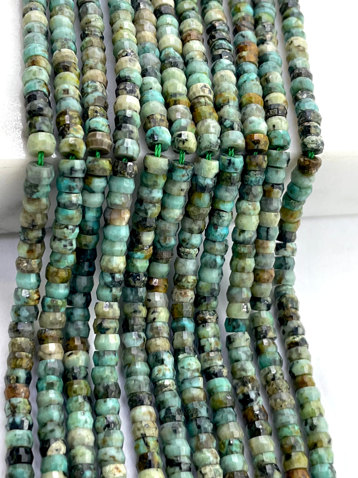 Multiple strands of faceted african turquoise made from natural stone, featuring a mix of turquoise, green, brown, and black colors, arranged vertically on a white background.