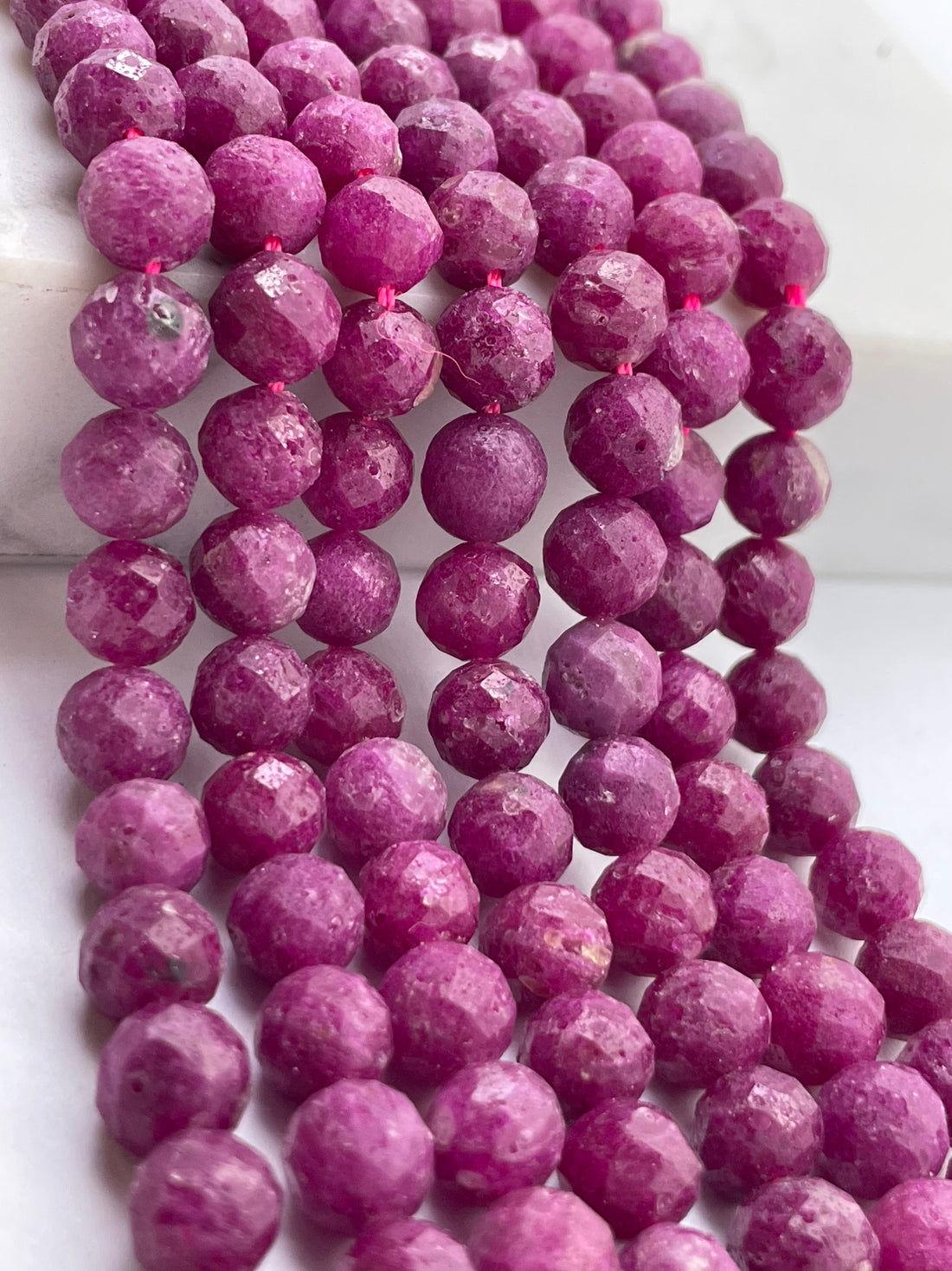 Faceted round ruby gemstone beads in a vibrant magenta-pink color, arranged in multiple strands on a white surface.