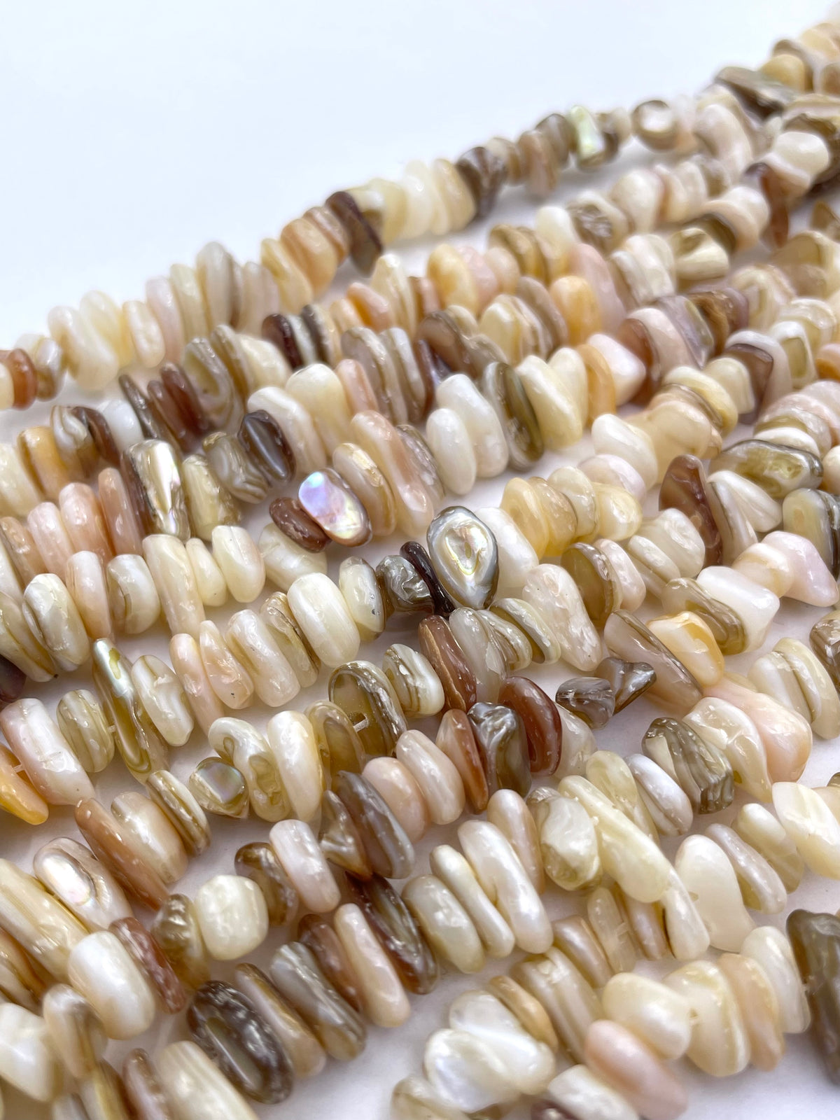 Close-up of multiple strands of small polished stone beads in mixed earthy tones including cream, beige, tan, brown, and some iridescent highlights, arranged on a white background.