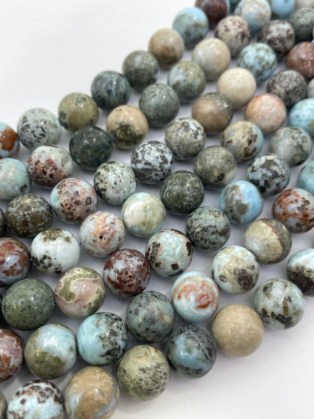 Round polished larimar gemstone beads in a marbled pattern of light blue, green, beige, brown, and white, arranged on a plain white background.
