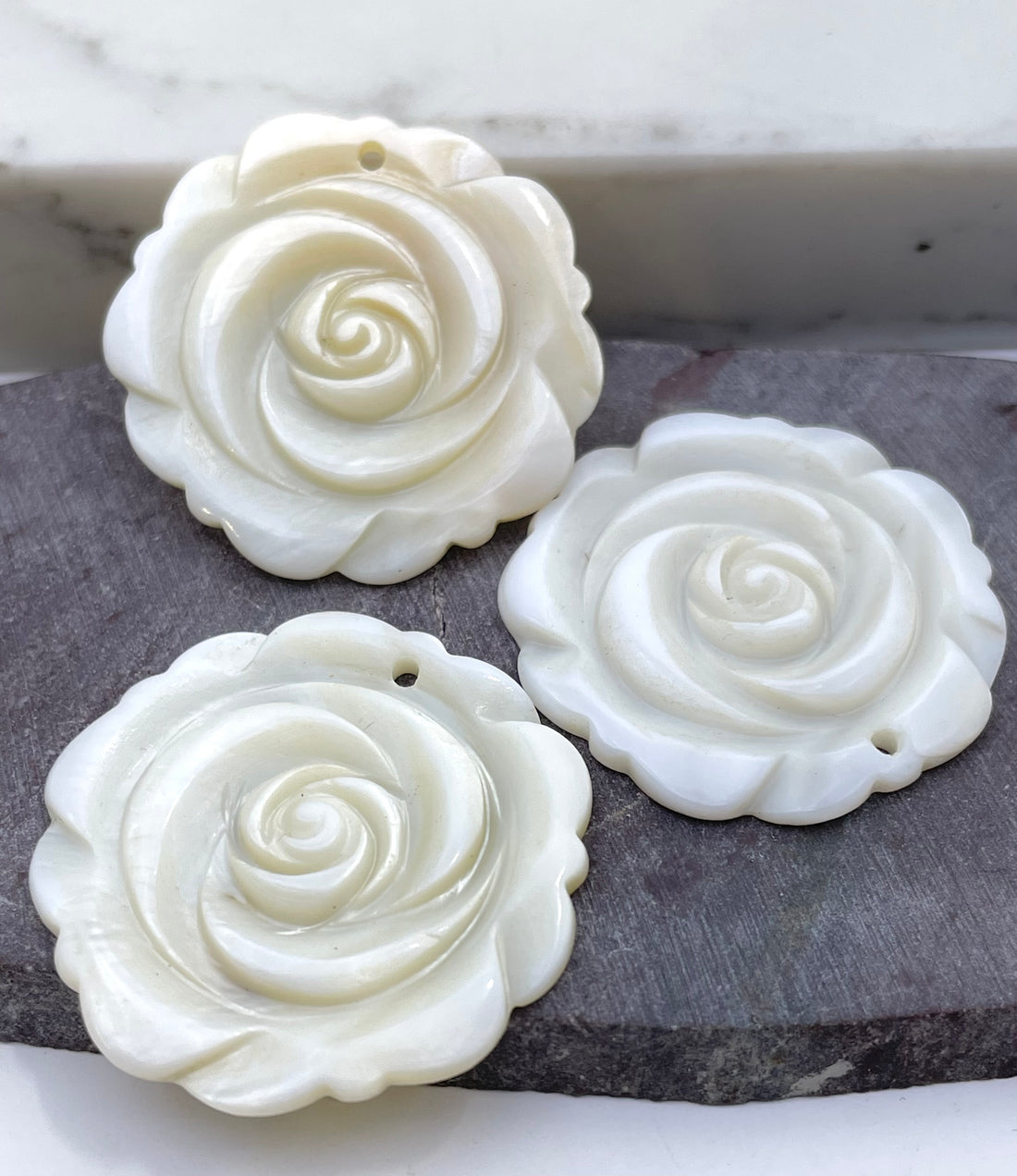 Three carved white rose-shaped jewelry pendants with a small hole at the top, shown with one held in a hand and two resting on a gray stone surface.