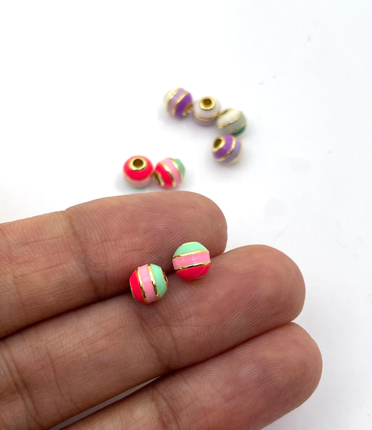 Two small striped brass beads held between fingers for scale, with several more beads scattered in the background on a white surface.