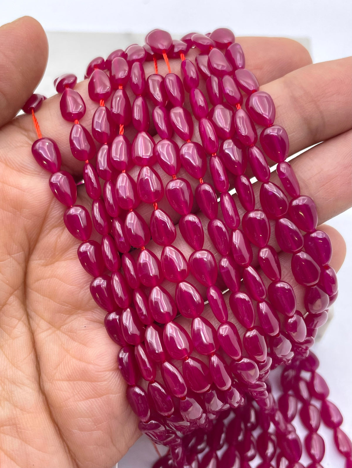 Multiple strands of glossy, teardrop-shaped lab ruby magenta beads held in a person's hand against a plain white background.