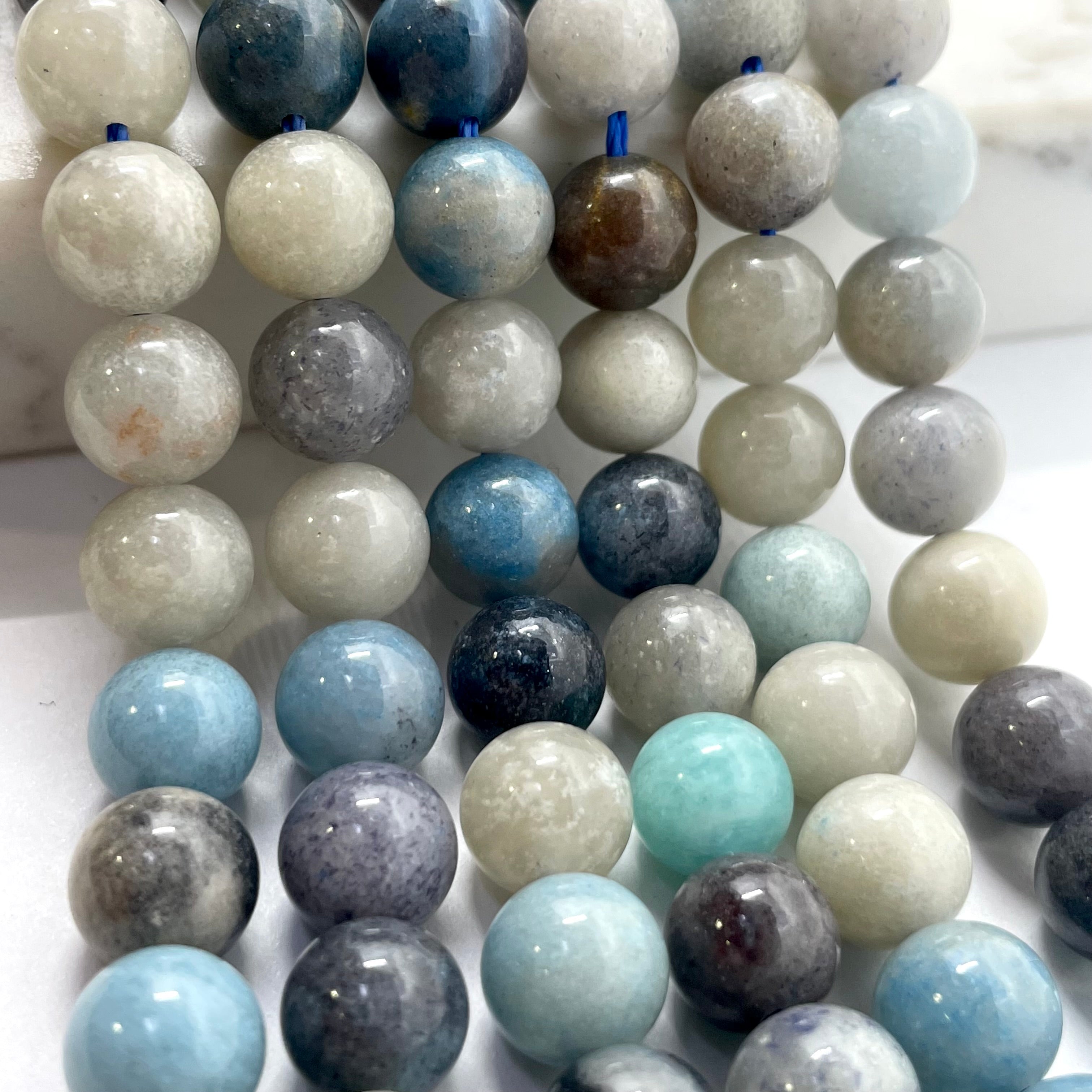 A close-up of multiple strands of polished round gemstone beads in assorted colors, including light blue, white, gray, navy, and marbled tones, arranged on a light surface.