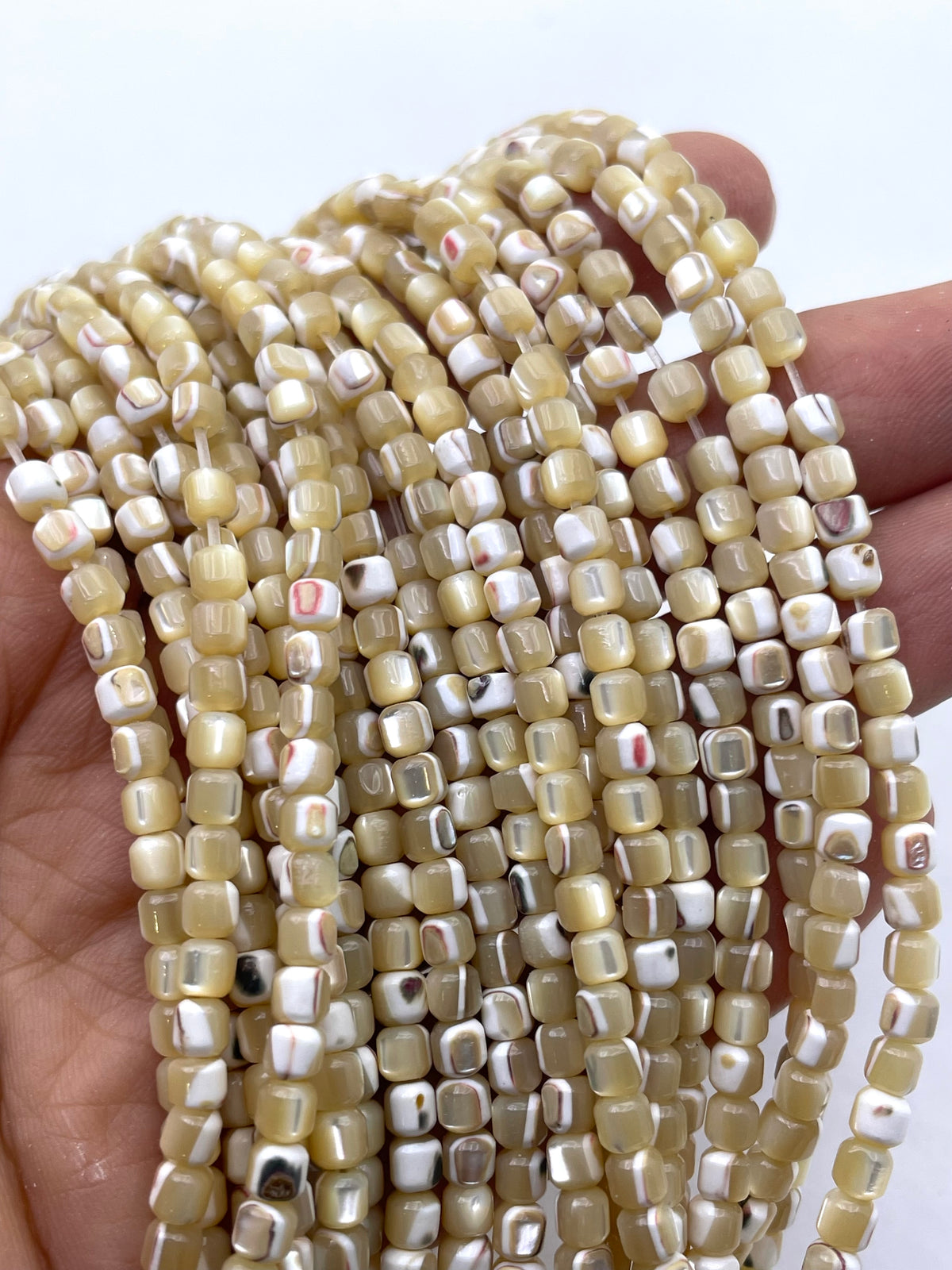 A close-up of multiple strands of small caramel mop square beads held in a hand. The beads are primarily a warm cream/beige color with white and black marbled or striped patterns running through them.