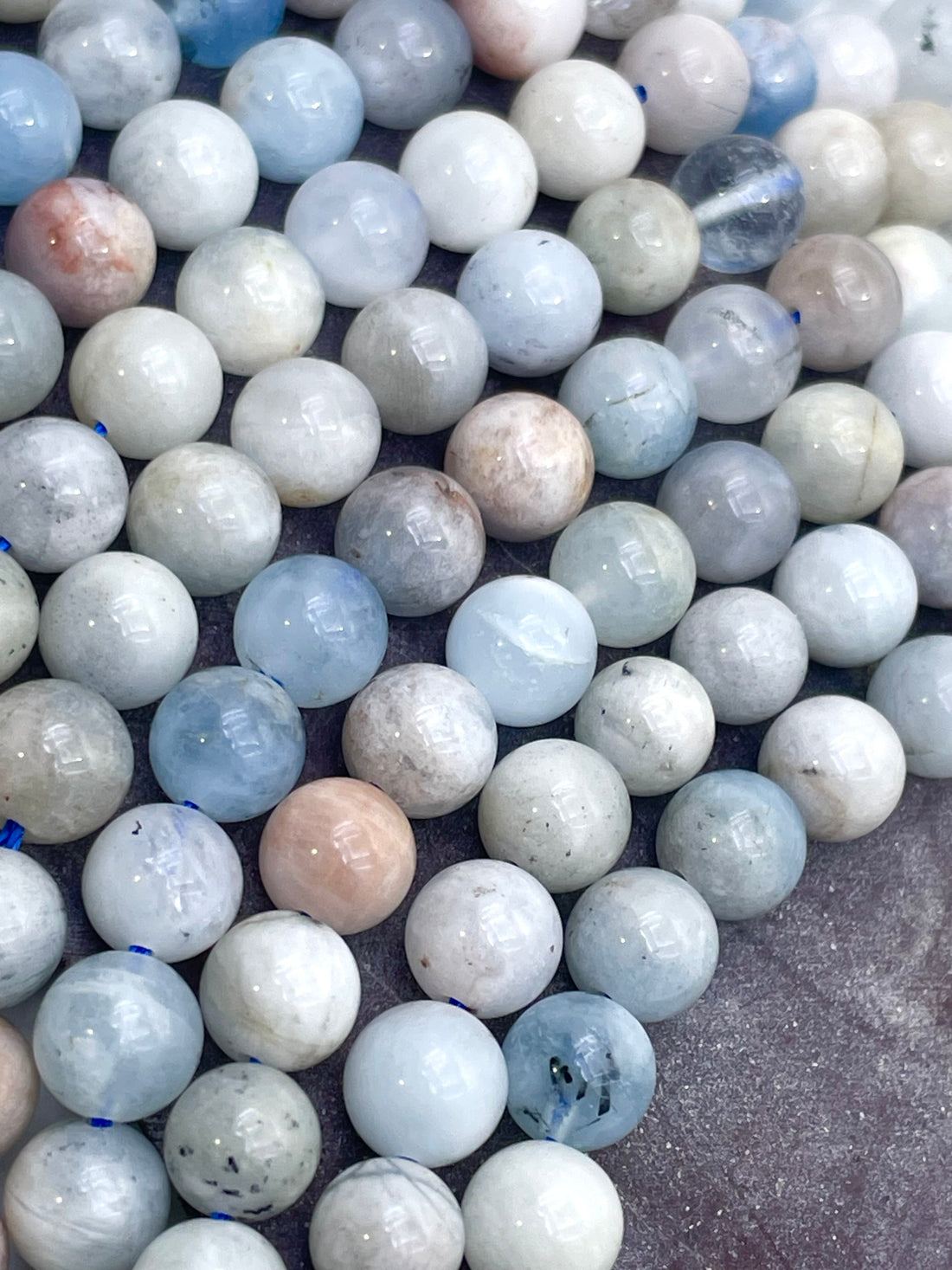 A close-up of polished round aquamarine gemstone beads in soft pastel shades of pale blue, white, gray, and faint peach, arranged on a textured gray surface.