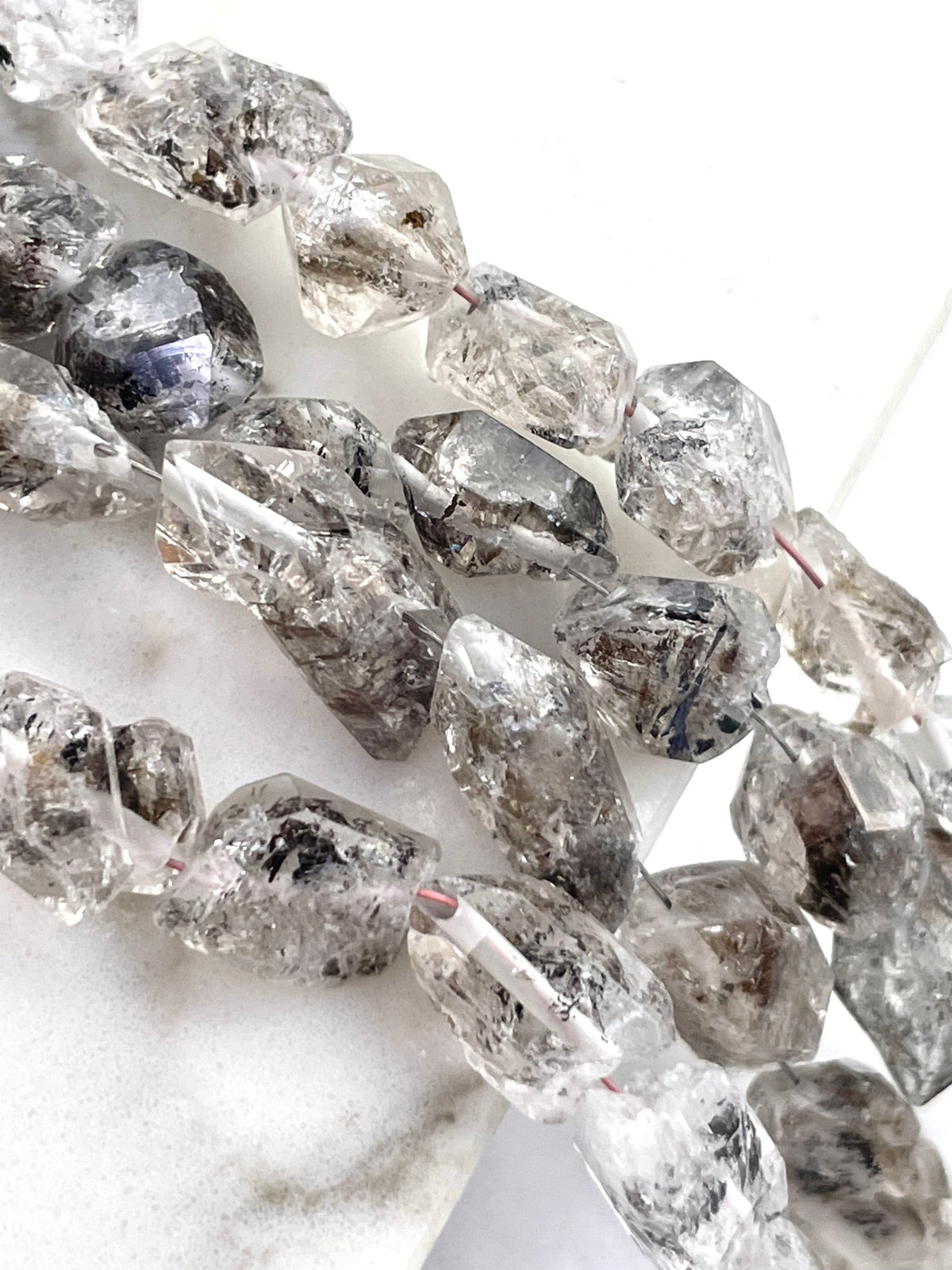 Herkimer diamond beads with natural inclusions, shown in a close-up diagonal arrangement on a white marble surface.