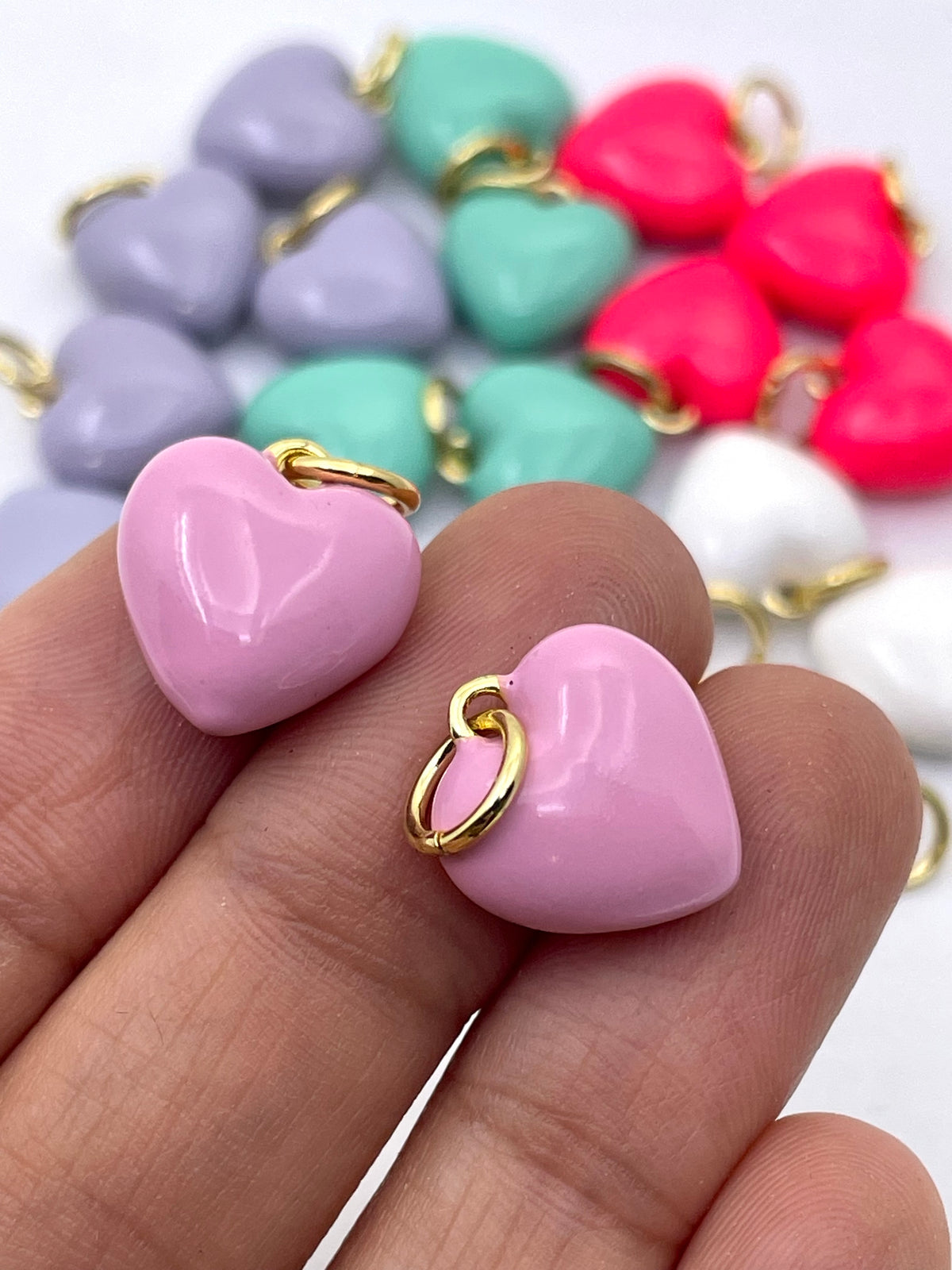 Two glossy pink heart-shaped charms with gold-tone loops, held on a person's fingers, with additional multicolor heart charms blurred in the background.