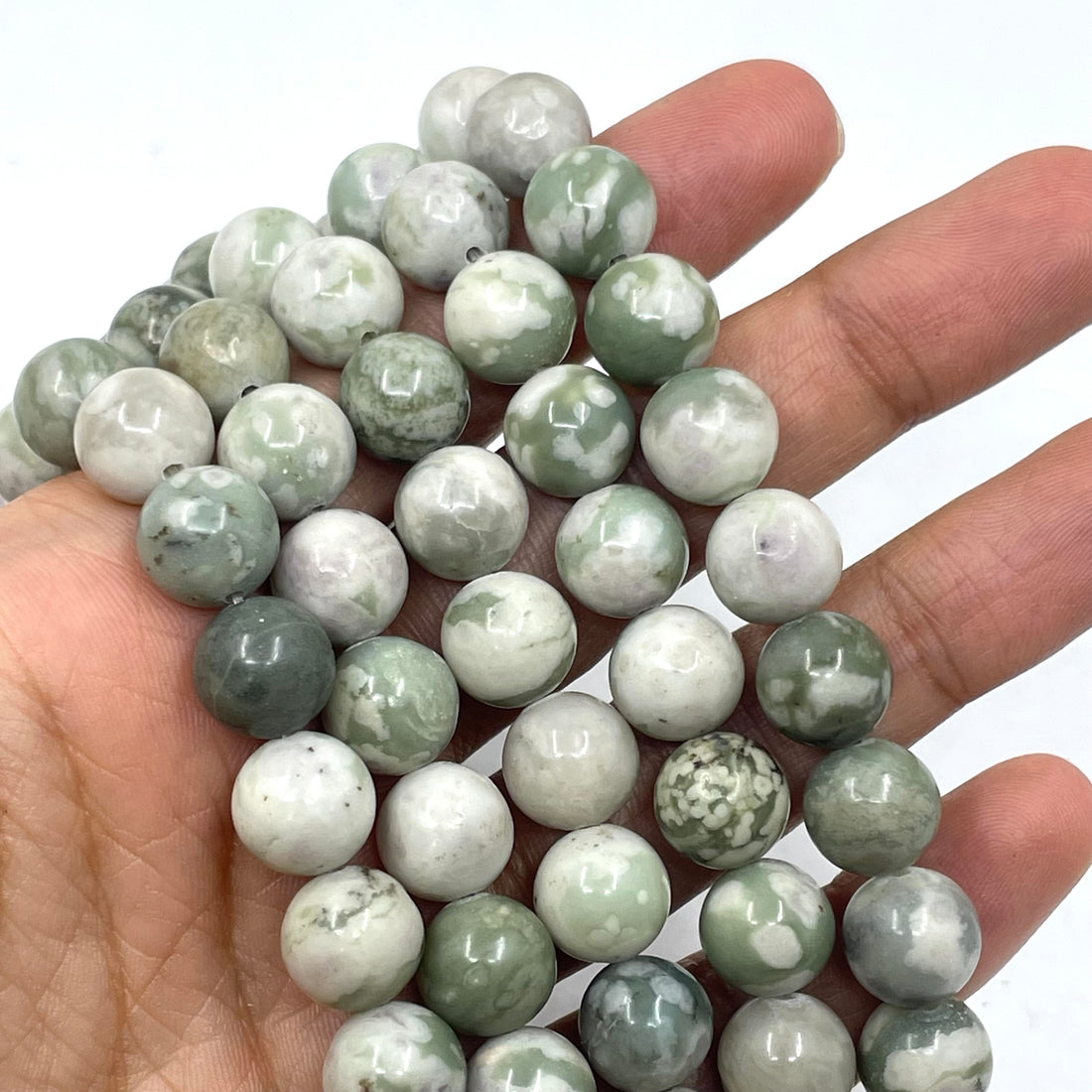 A close-up of polished round peace jade gemstone beads or beads arranged in rows on a white background. The beads are predominantly light gray and white with natural green and beige marbling.