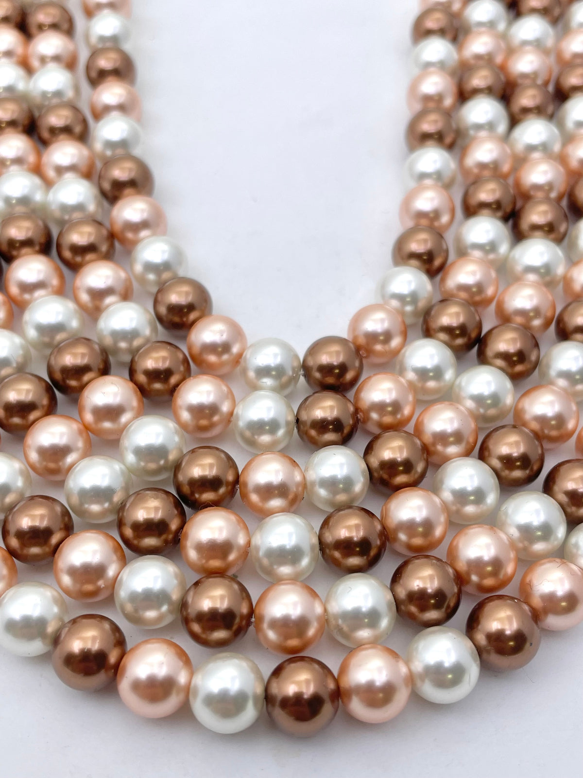 Multiple strands of shell pearl beads in rose gold, champagne, and white, arranged in a layered V-shape on a plain white background.