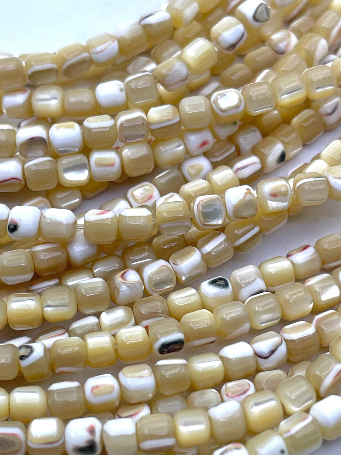 Close-up of multiple strands of small caramel mother of pearl square beads with a glossy finish, featuring a mix of cream, beige, white, and subtle black and red marbled patterns.