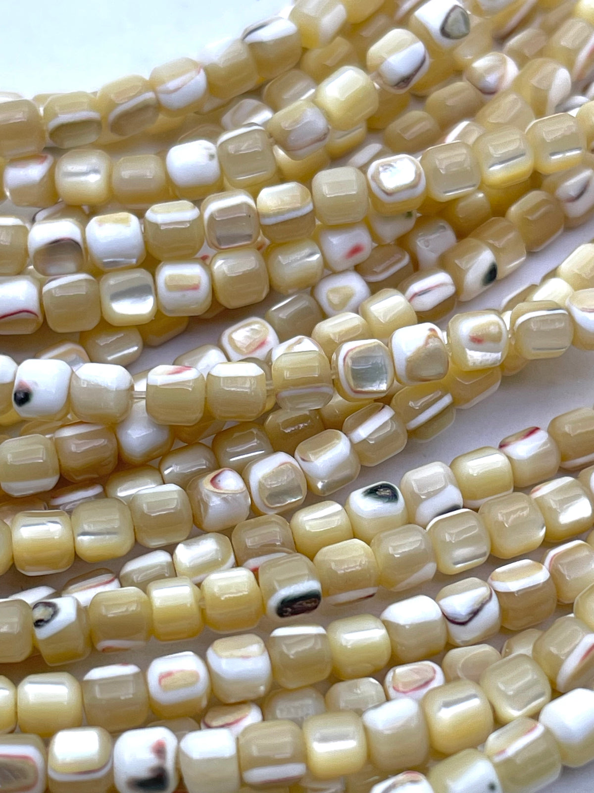 Close-up of multiple strands of small caramel mother of pearl square beads with a glossy finish, featuring a mix of cream, beige, white, and subtle black and red marbled patterns.