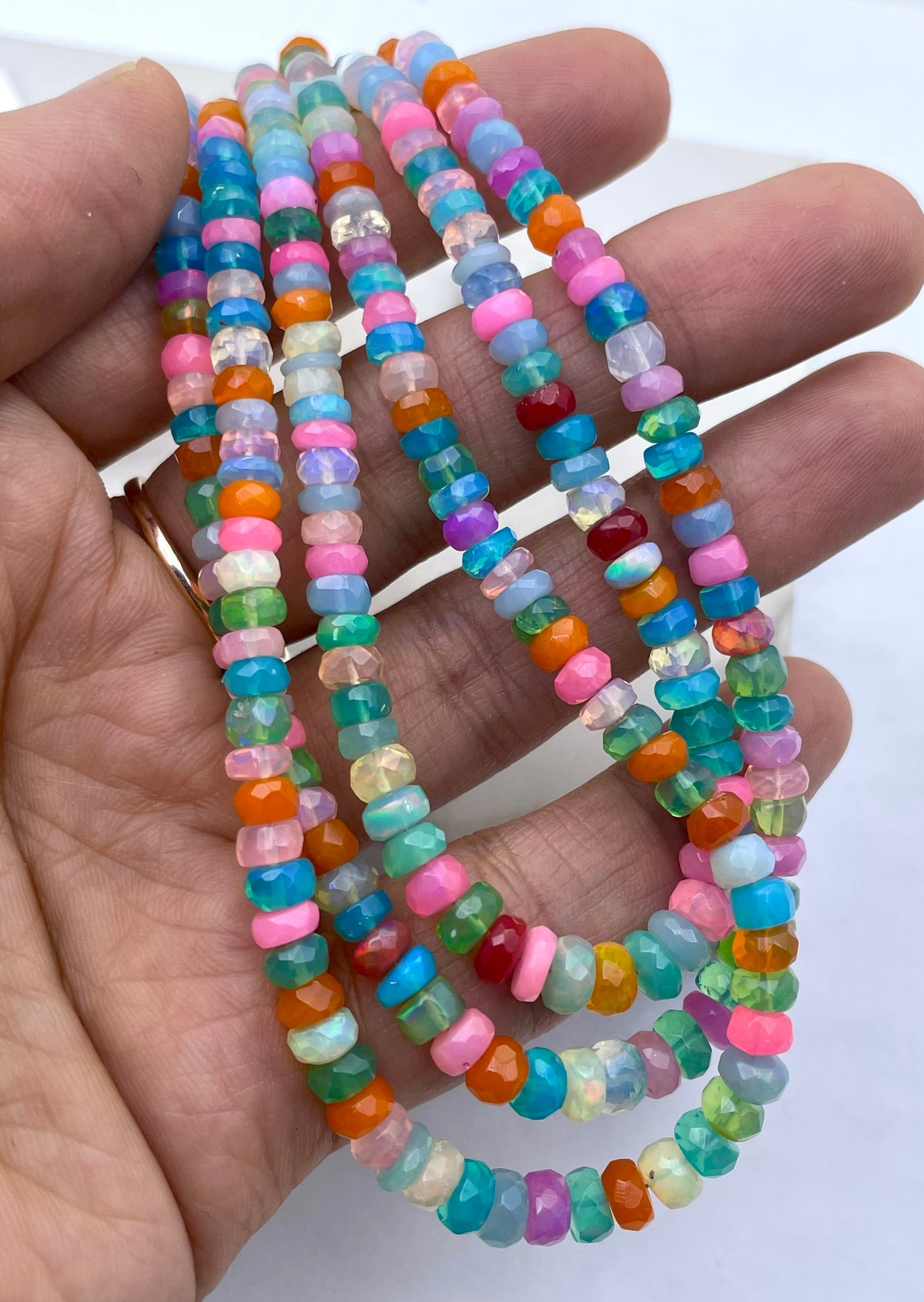 A close-up of a hand holding three strands of colorful faceted ethopian opal gemstone beads, including pink, blue, green, orange, purple, and white beads.