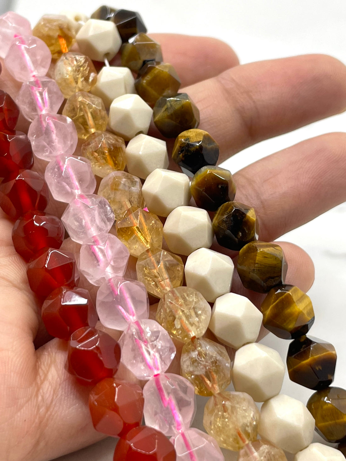 Close-up of faceted gemstone beads draped together on a white surface, including carnelian, rose quartz, citrine, fossil stone and tiger eye