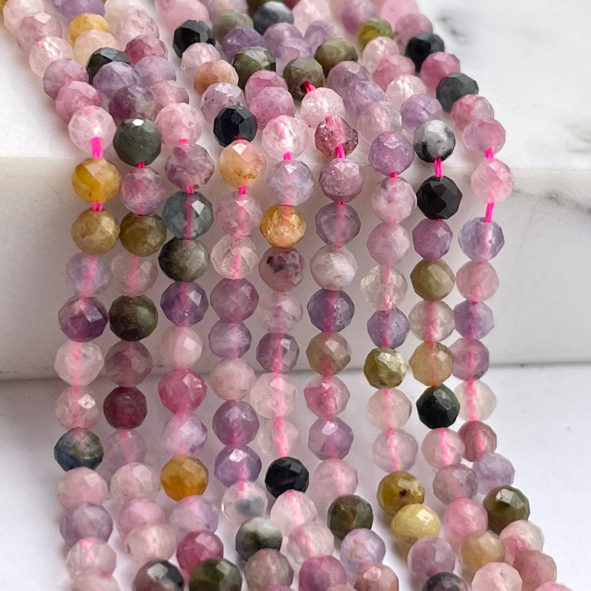 Multiple strands of faceted round mix tourmaline gemstone beads strung on pink thread, displayed on a white marble surface. The beads feature a mix of pink, purple, green, yellow, and black tones.