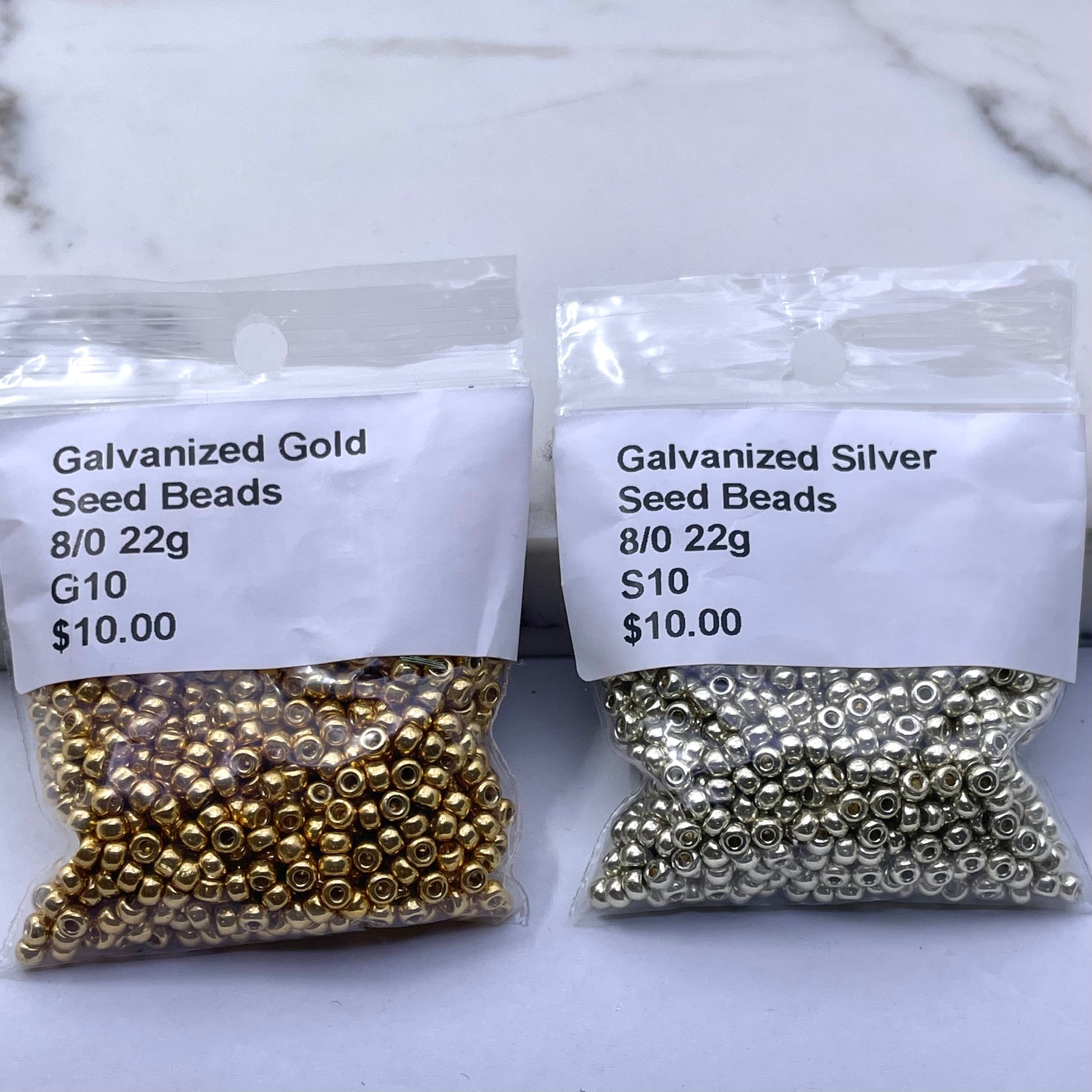Two clear plastic bags of seed beads on a light marble-like surface. The left bag contains gold-colored beads with a white label reading 'Galvanized Gold Seed Beads 8/0 22g G10 $10.