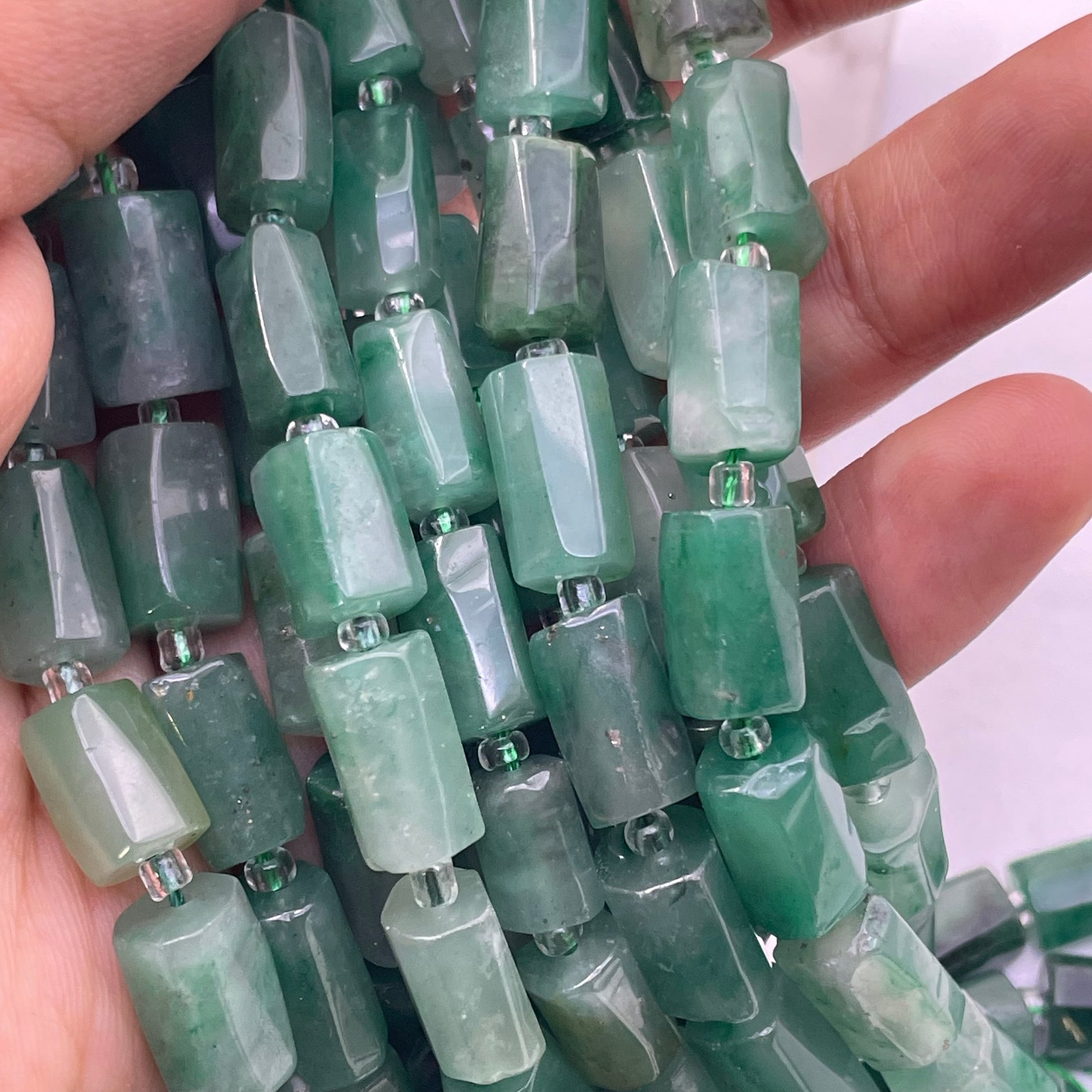 A close-up of a hand holding multiple strands of faceted african green jade beads, showing their translucent green color and polished edges.