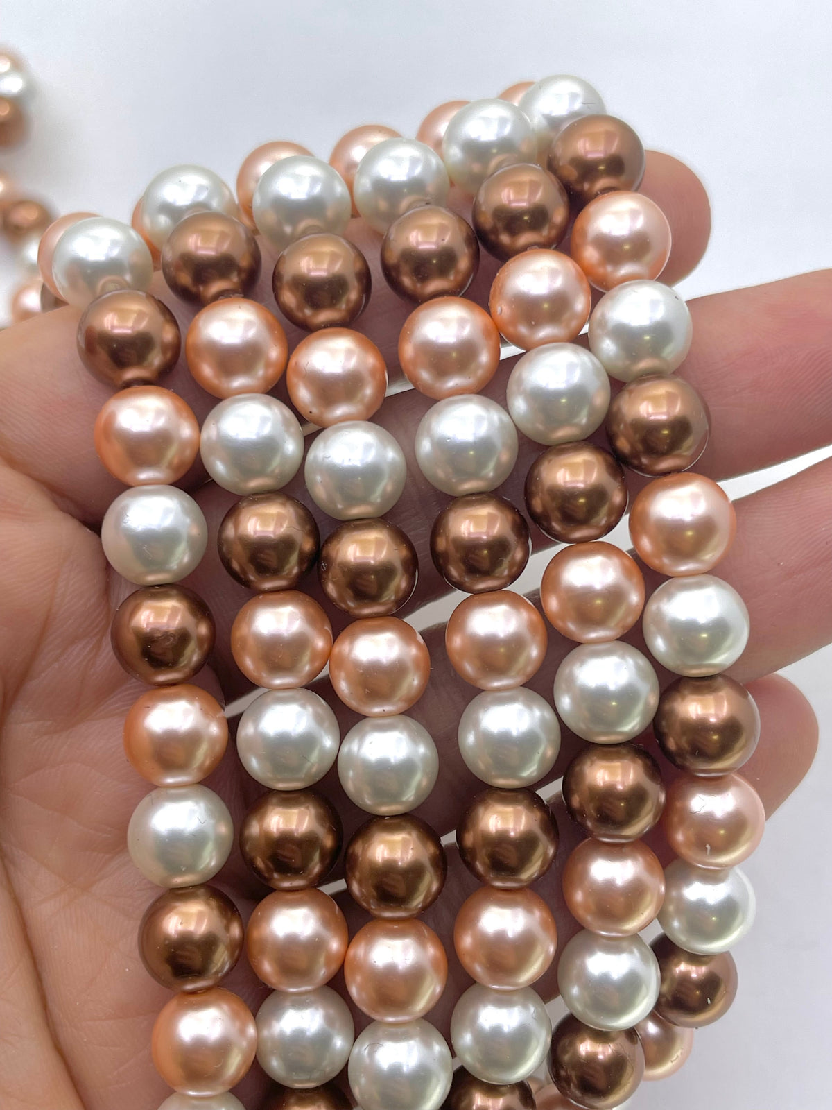 A hand holding multiple strands of round shell pearls in mixed colors including white, cream, and metallic copper and rose-gold tones, set against a plain white background.