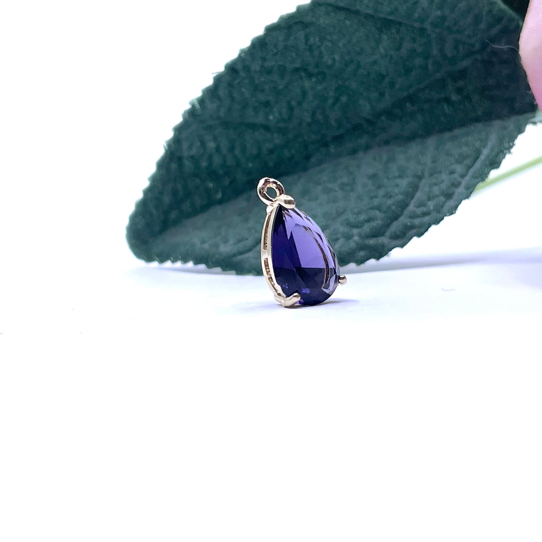 Five teardrop-shaped faceted crystal pendants in light blue, dark purple, clear, red, and green, each with a gold-tone loop at the top, arranged on a white surface with blurred green leaves and a pink flower in the background.