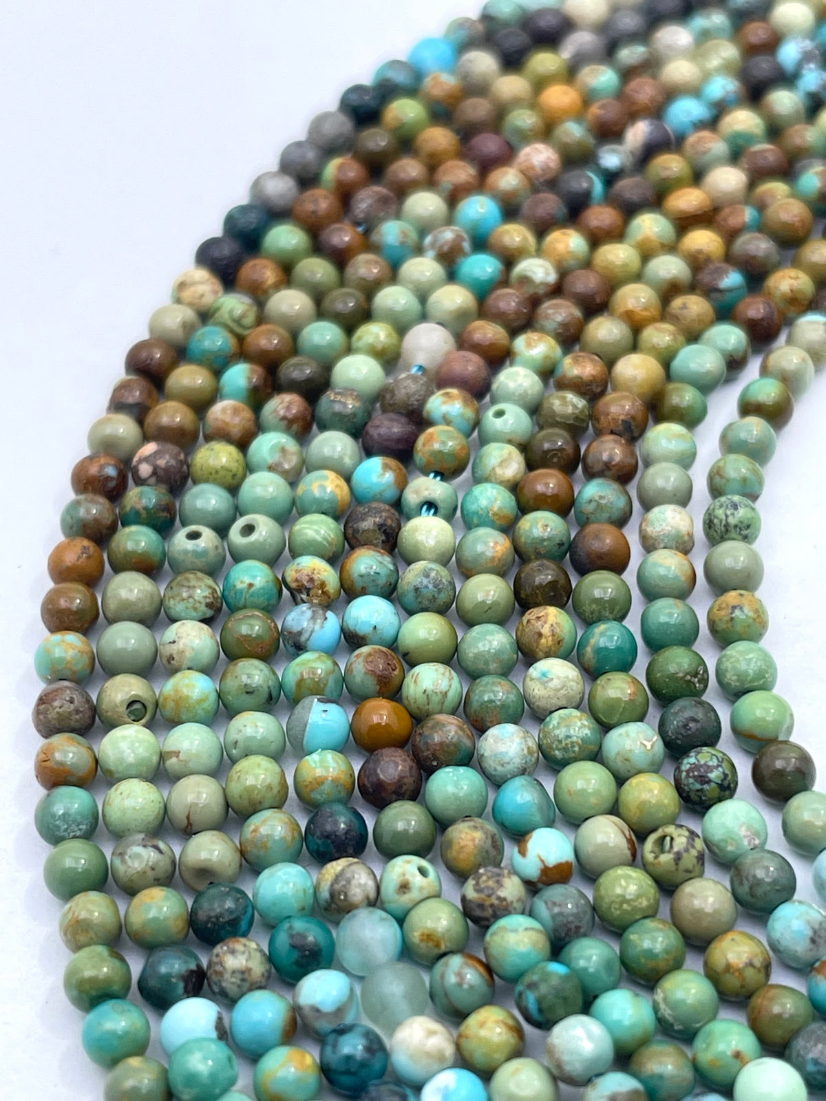 Curved strands of small round turquoise stone beads in various earthy green, blue, brown, and beige tones on a white background.